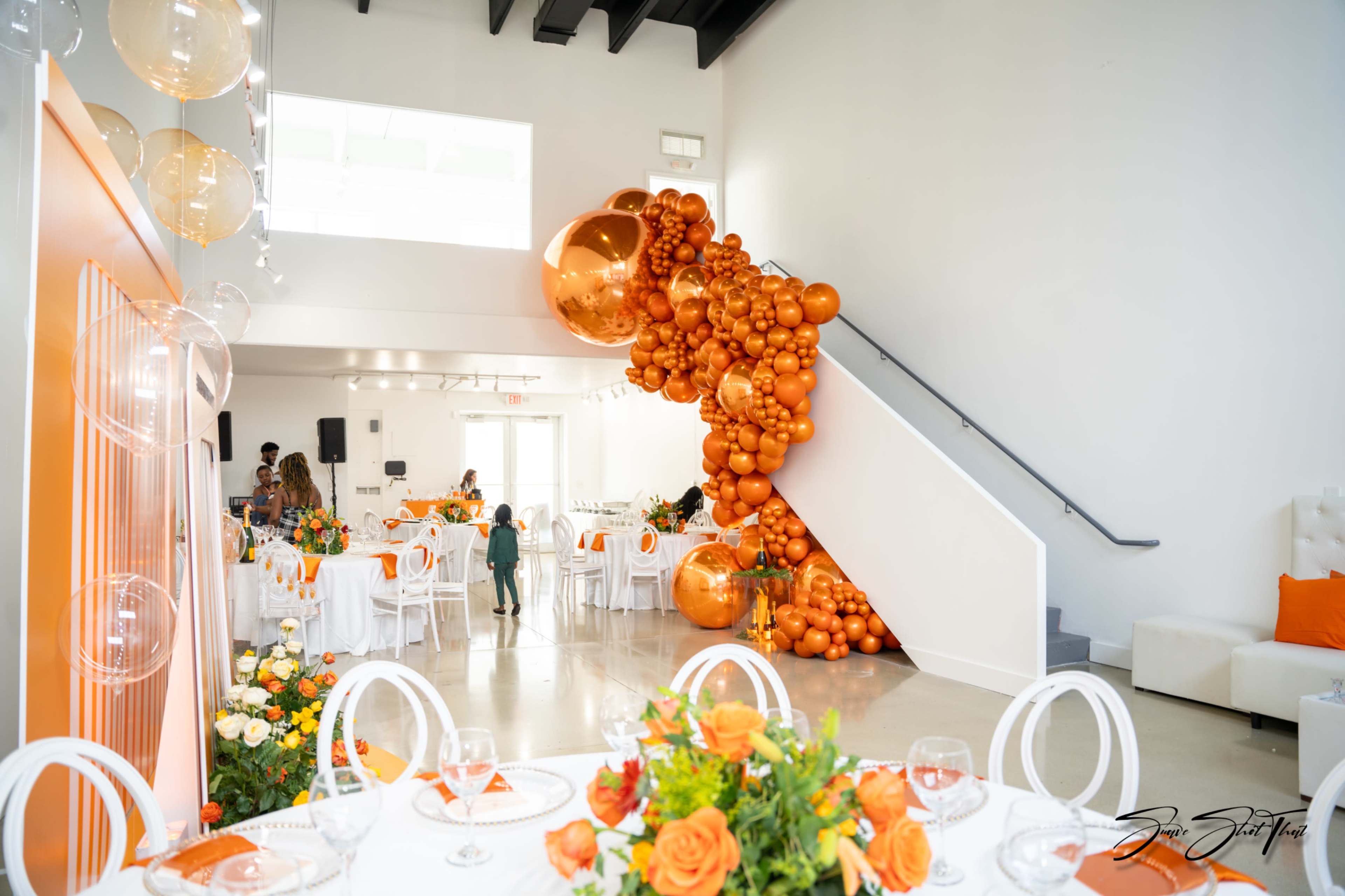 The image shows a brightly decorated event space with orange balloon installations and elegantly set tables featuring floral centerpieces.