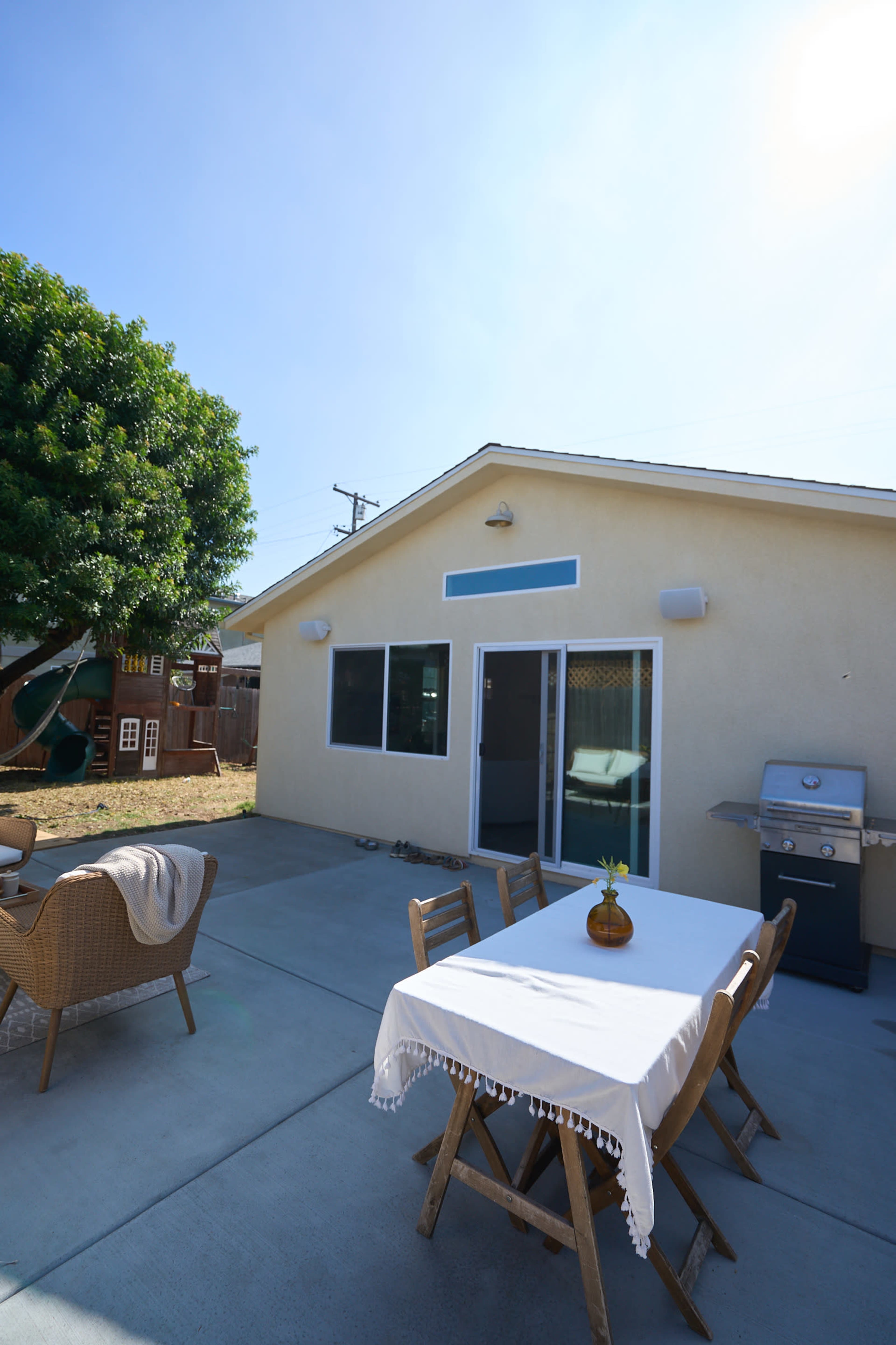 The image shows a backyard patio area with a table and chairs, a grill, and a small playhouse in the background.
