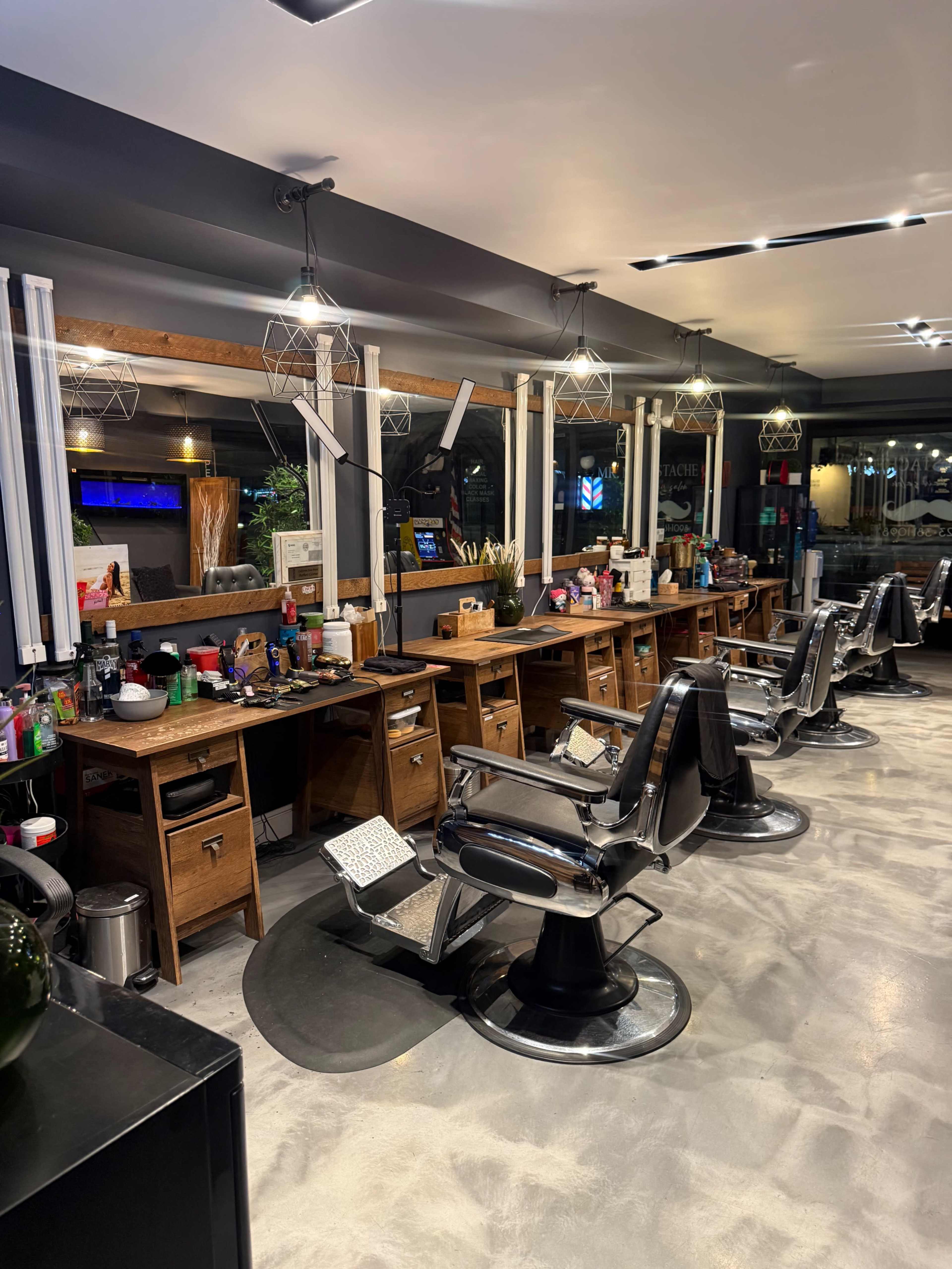 The image shows a modern barber shop interior with several barber chairs, wooden workstations, and contemporary lighting fixtures.