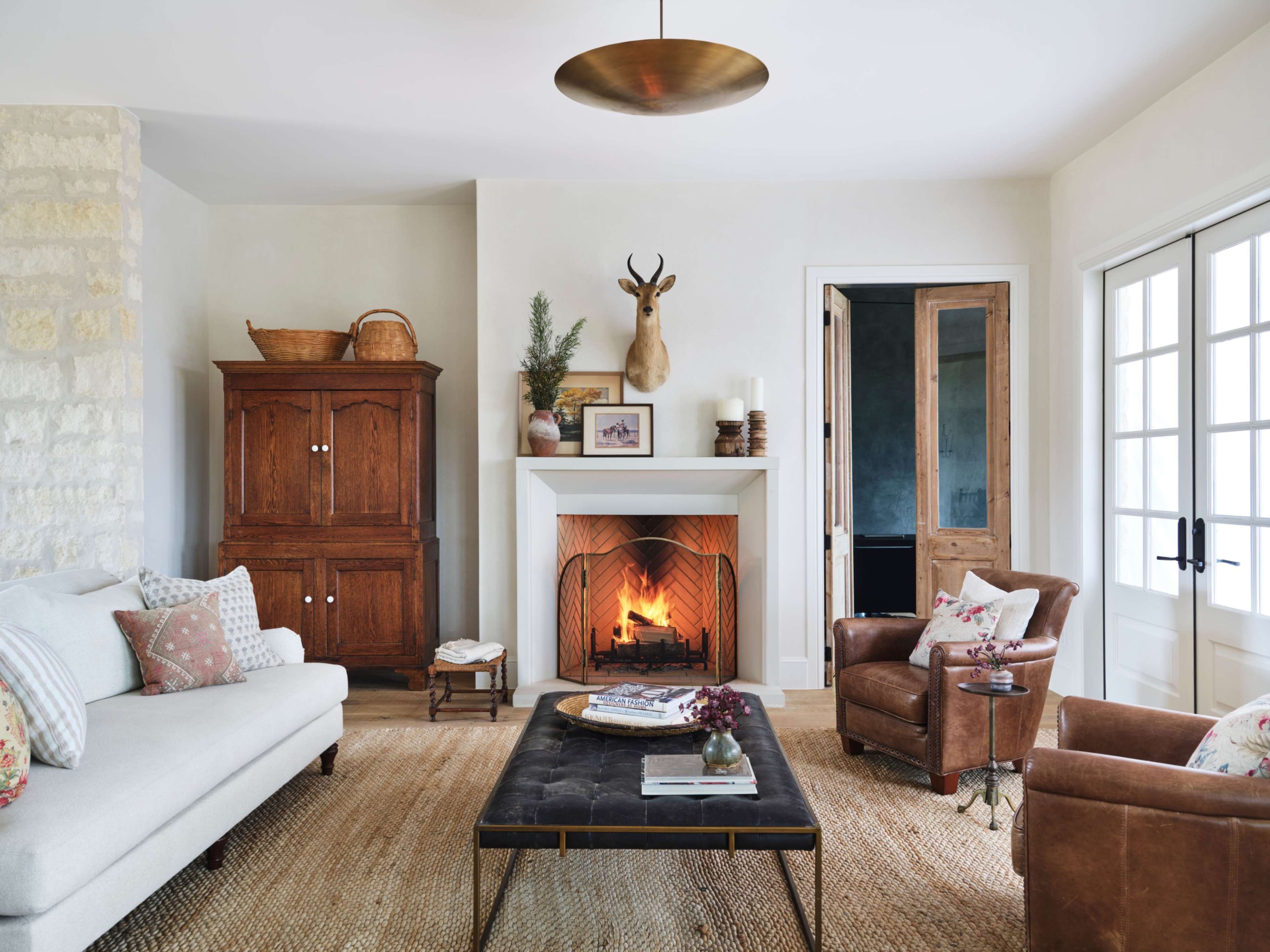 The living room features a fireplace with a mounted deer head above it, surrounded by a light-colored sofa, leather chairs, and a coffee table on a woven rug.