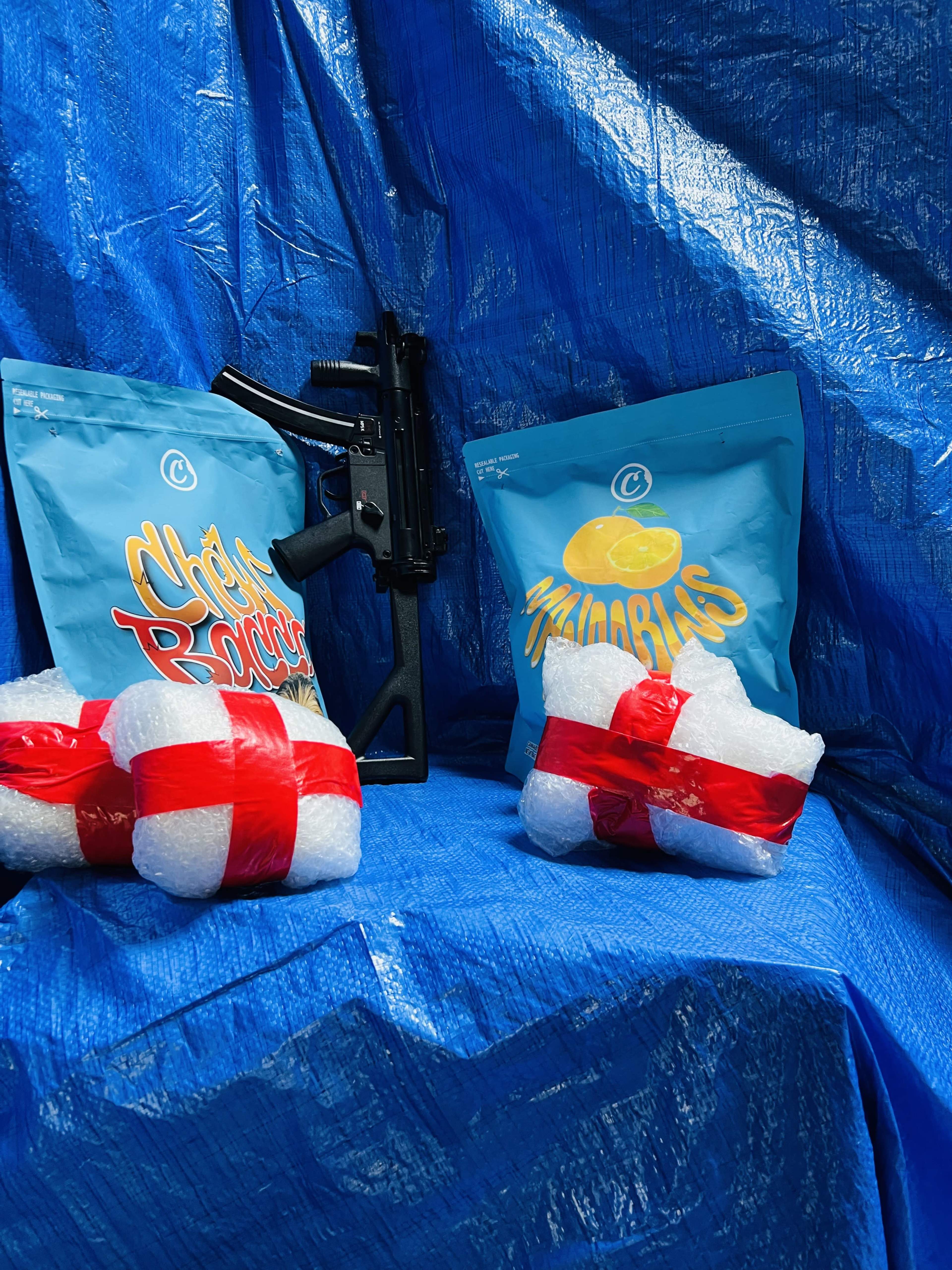 The image shows a black firearm beside two bags of snacks wrapped with red ribbons, placed on a blue tarp.