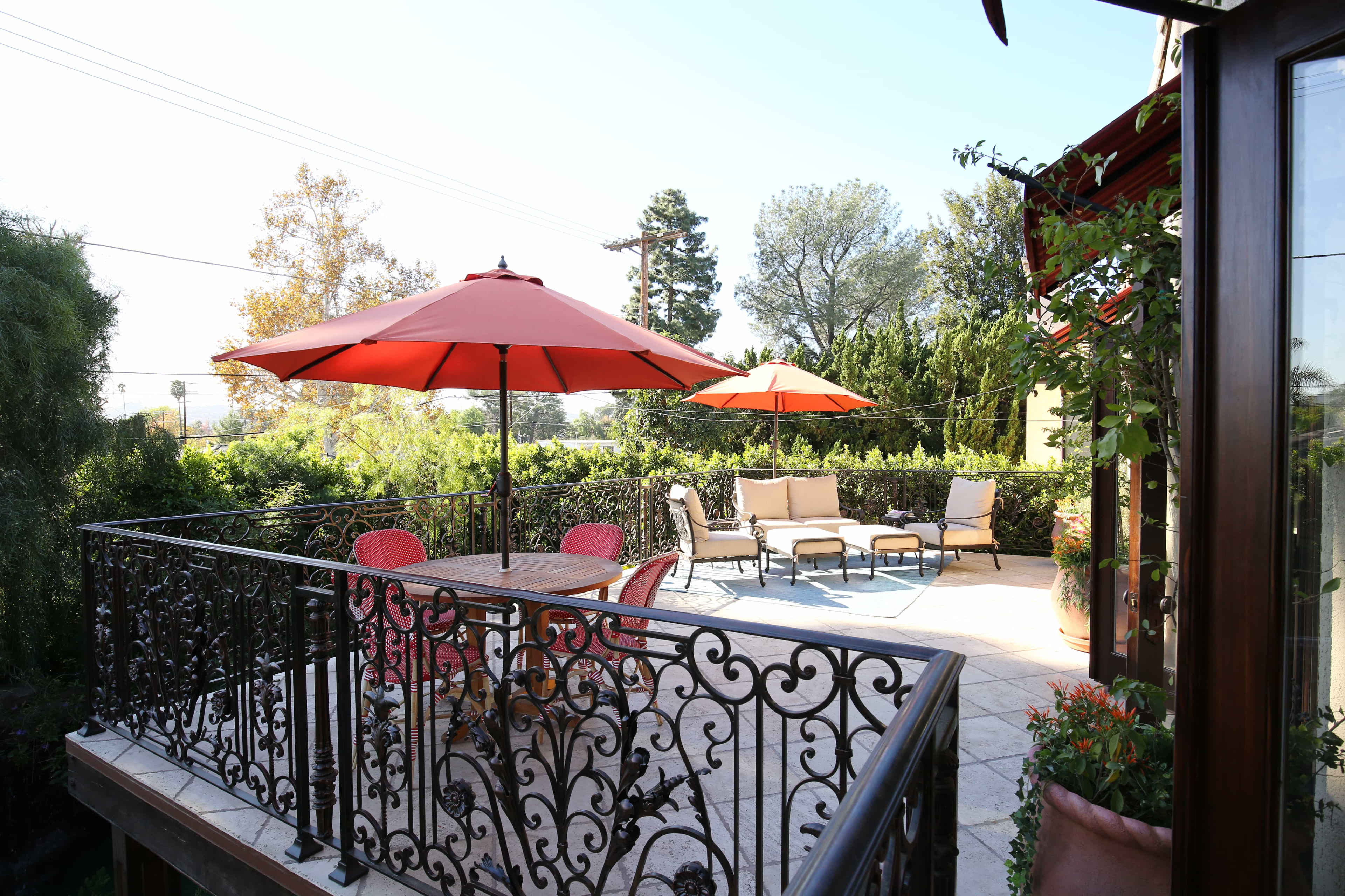 A patio features a wooden table surrounded by red chairs and two large red umbrellas overlooking a landscaped yard.