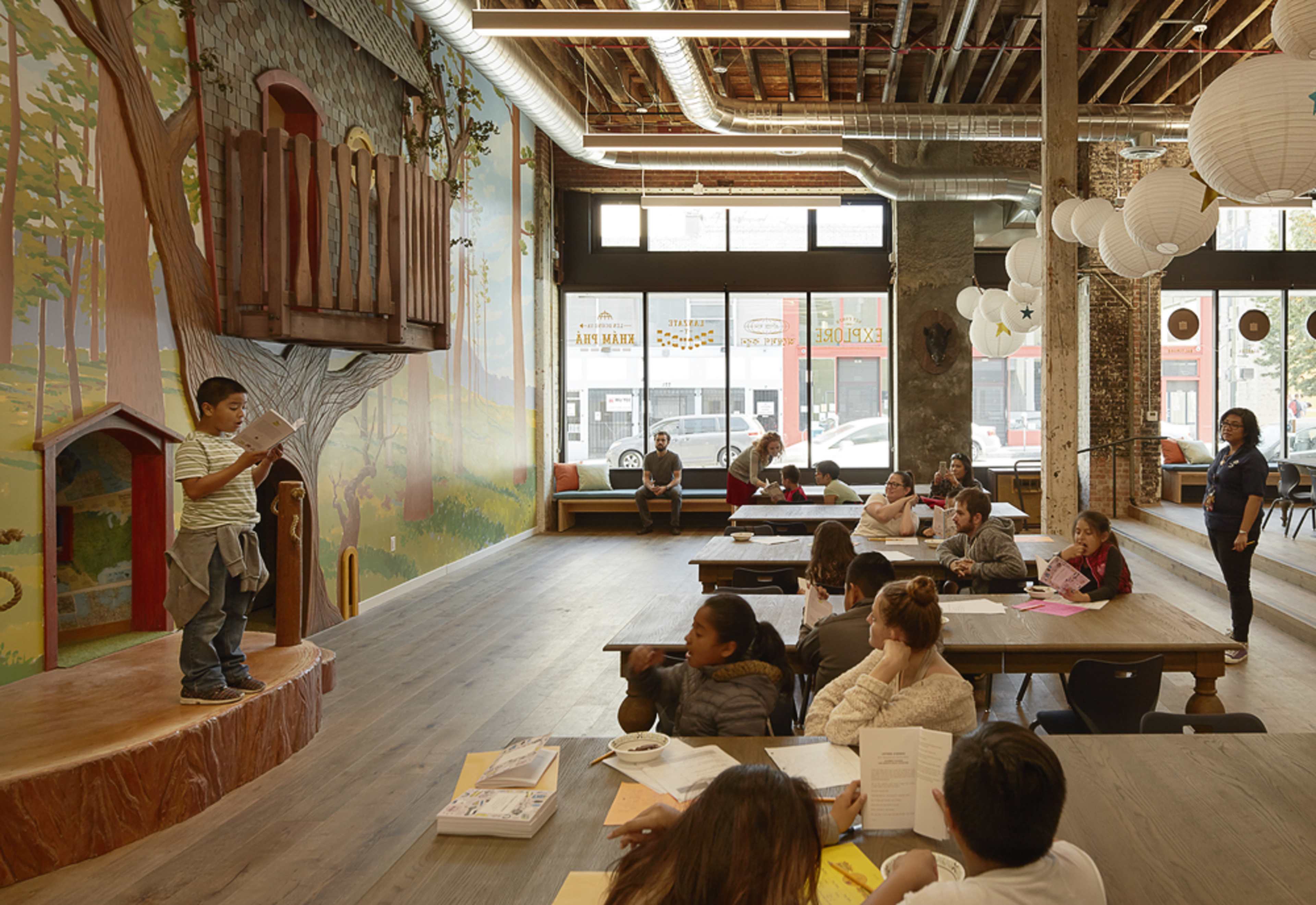 A child stands on a small stage reading aloud to a group of seated classmates in a brightly lit classroom decorated with a tree mural.
