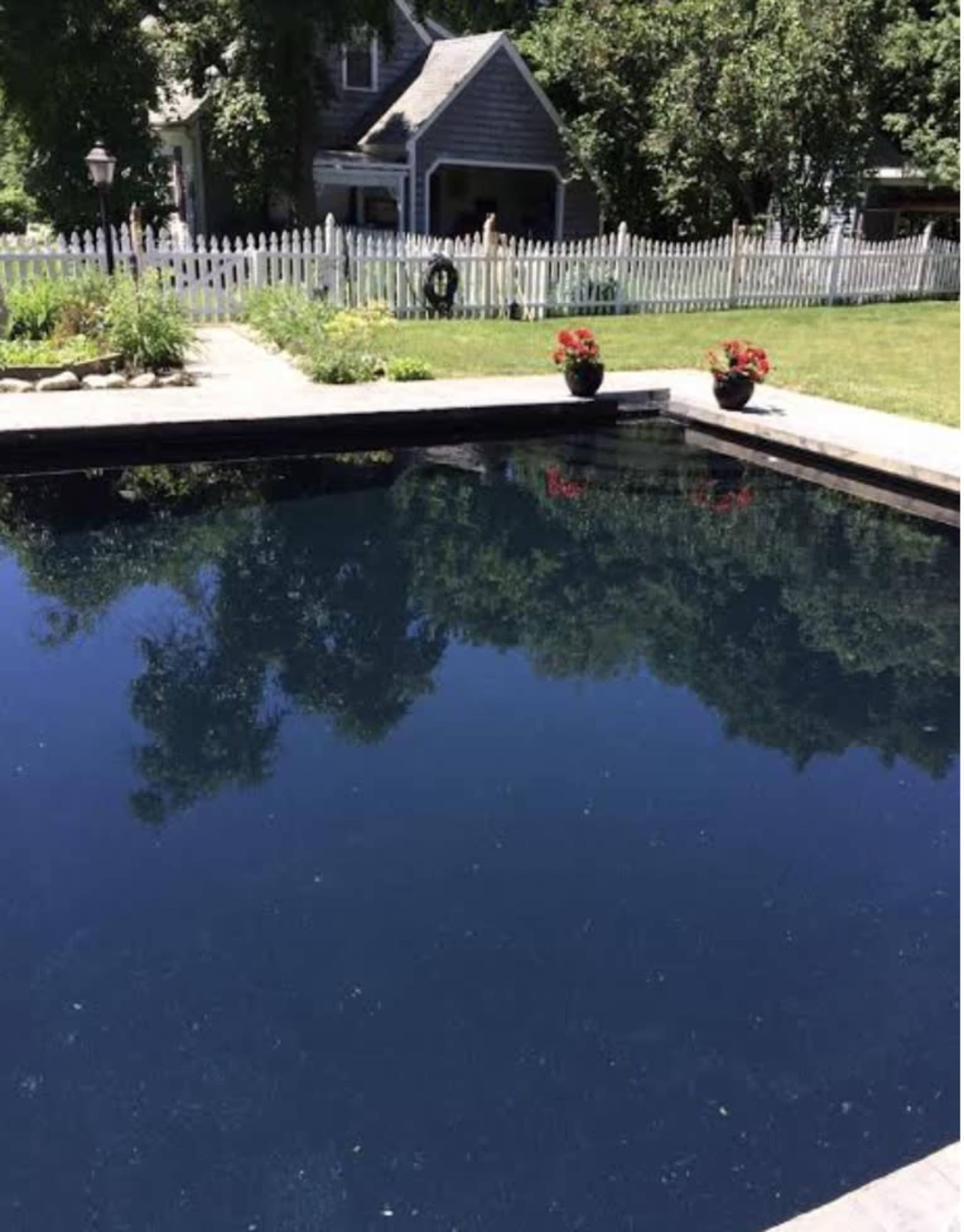 A dark, still swimming pool surrounded by a grassy area and a white picket fence, with a house visible in the background.