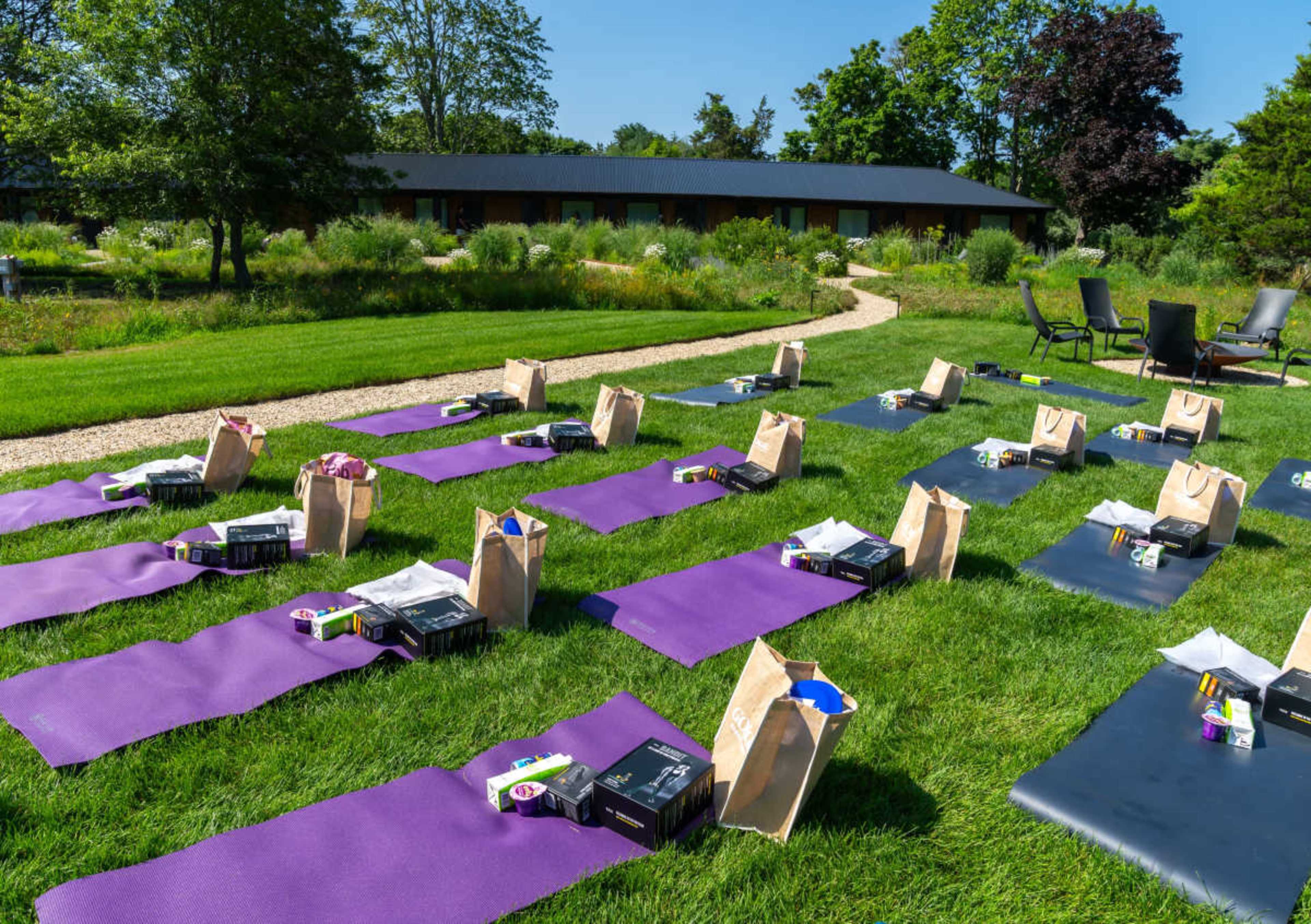 The image shows a series of yoga mats arranged on a grassy area, each accompanied by gift bags, with a backdrop of a building and trees.
