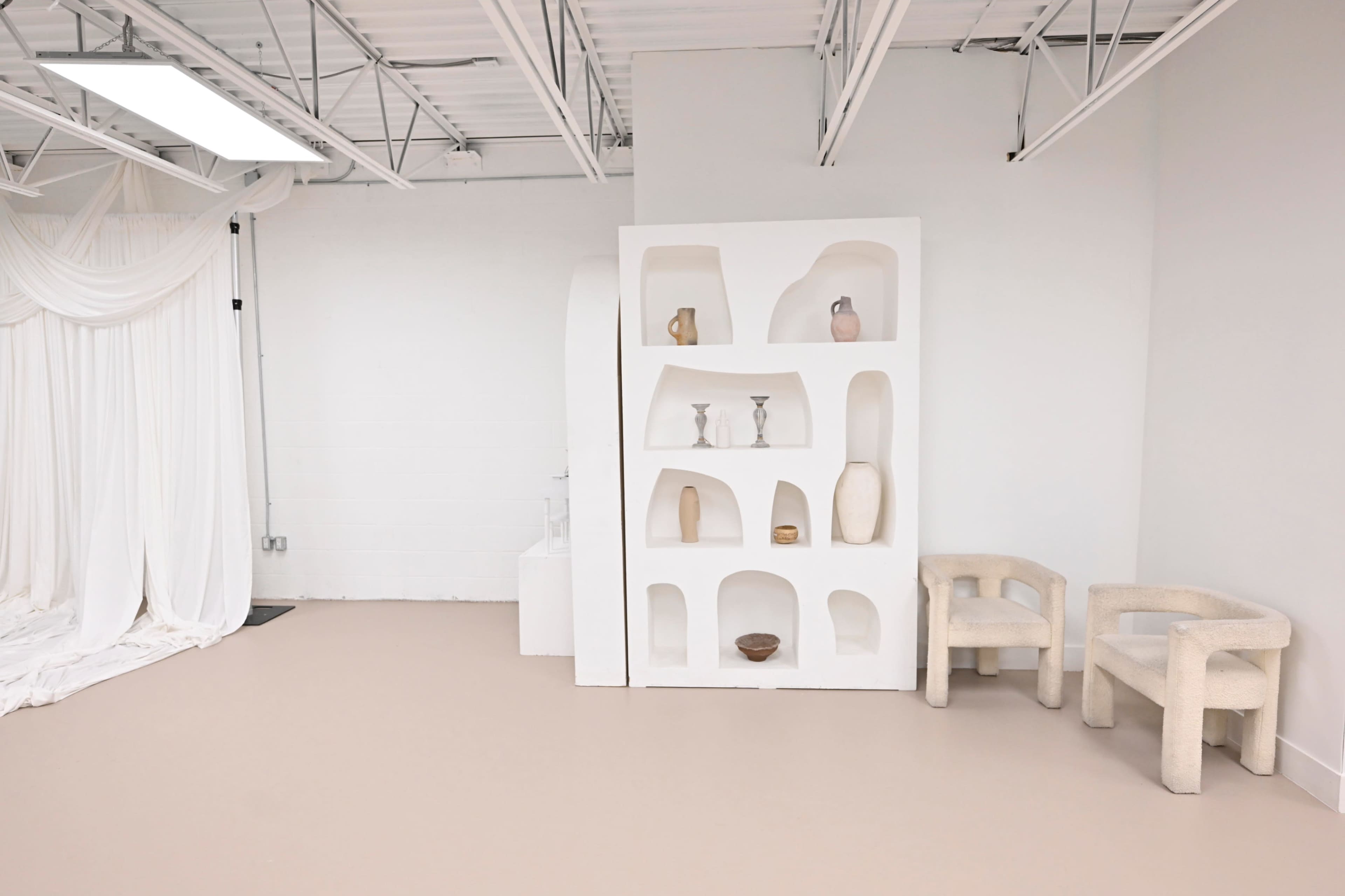 The image shows a minimalist interior space featuring a white shelving unit with various decorative items and a pair of rounded, sculptural chairs.