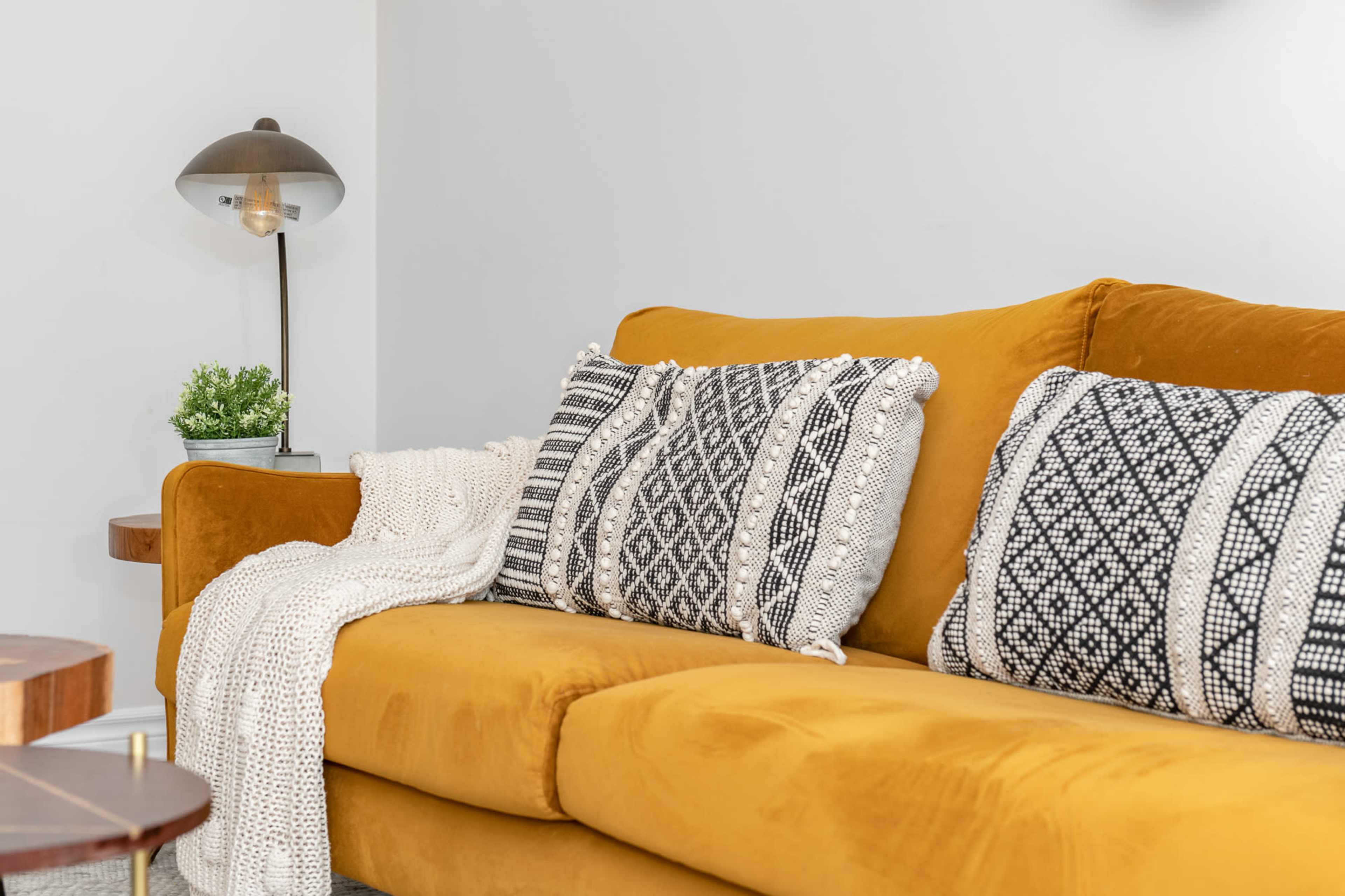 A mustard yellow couch with patterned black and white pillows is draped with a white knit blanket, positioned next to a small side table and a lamp.