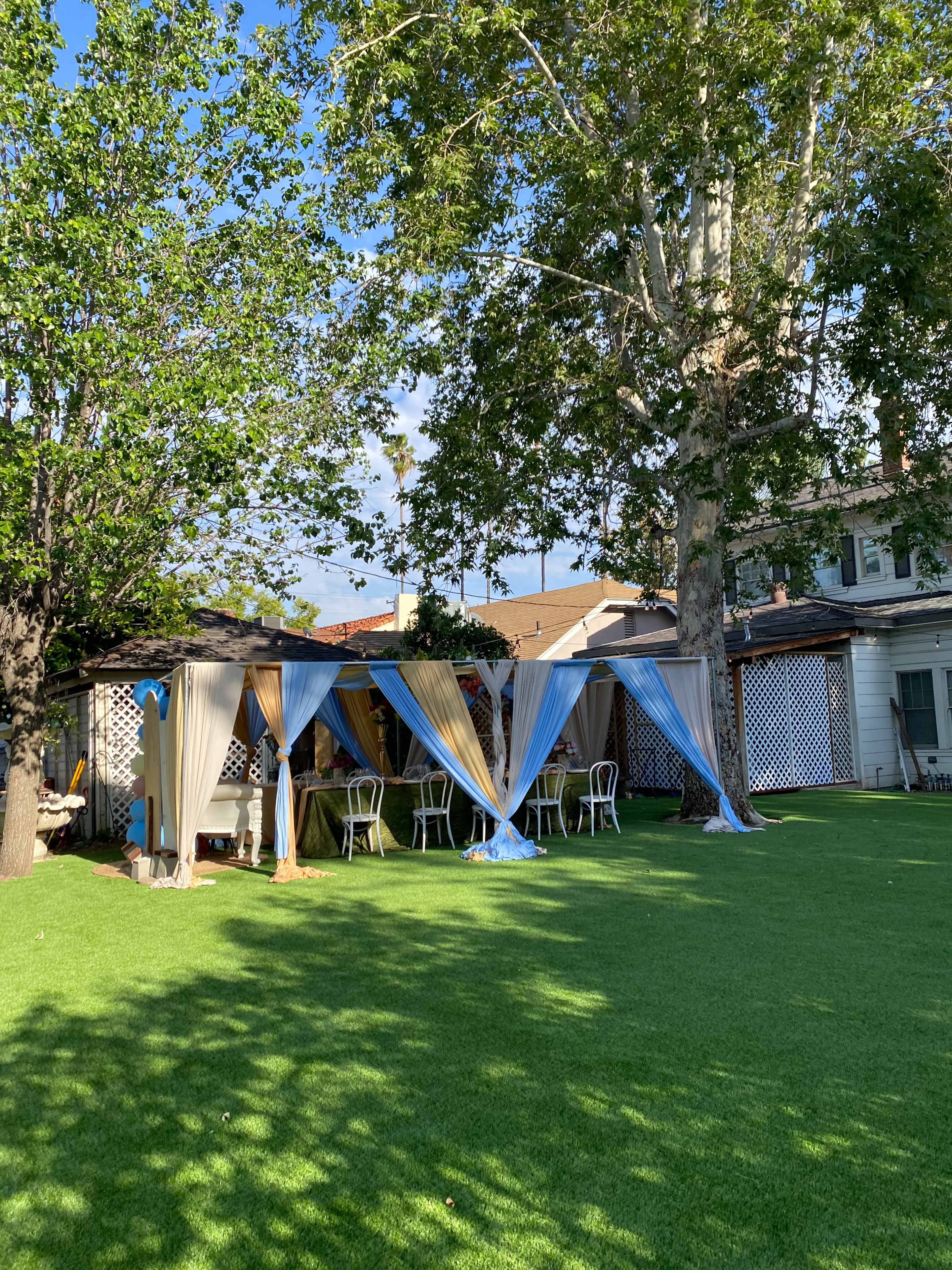 A secluded outdoor area with canopies draped in blue and beige fabric, surrounded by green grass and chairs under a large tree.