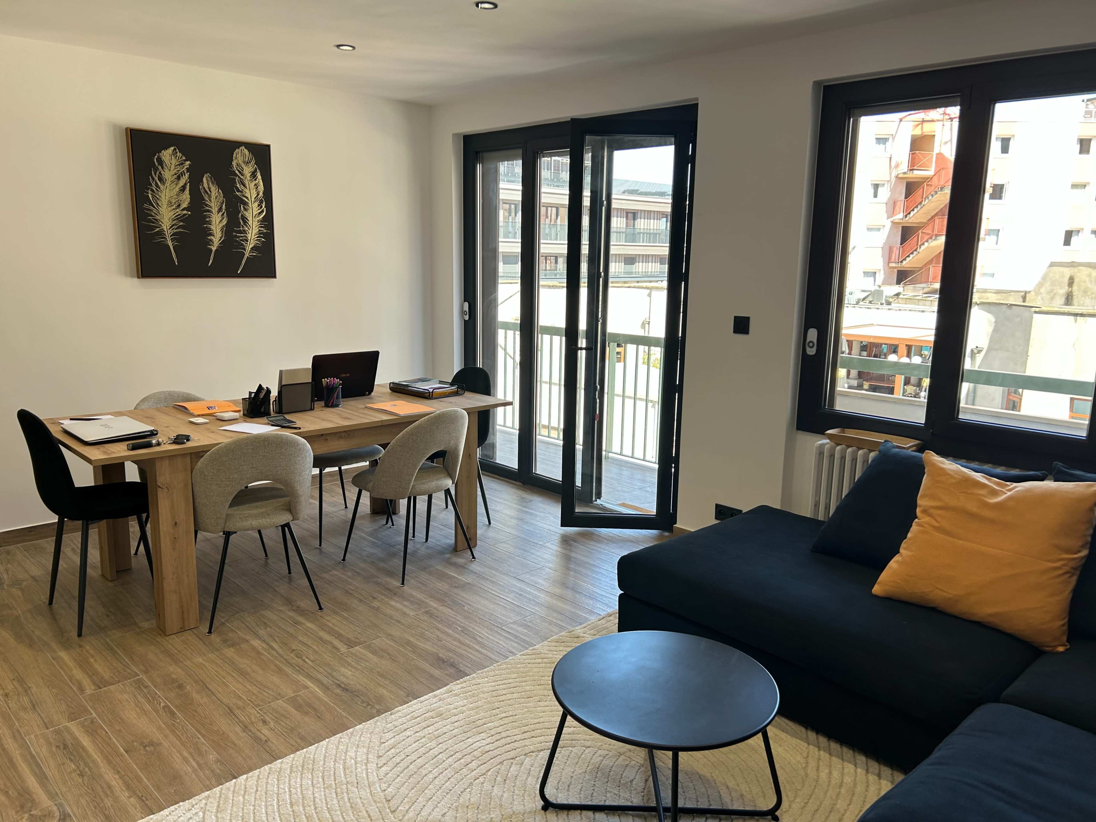The image shows a modern living area with a dining table, chairs, a dark sofa with a decorative pillow, and large windows leading to a balcony.