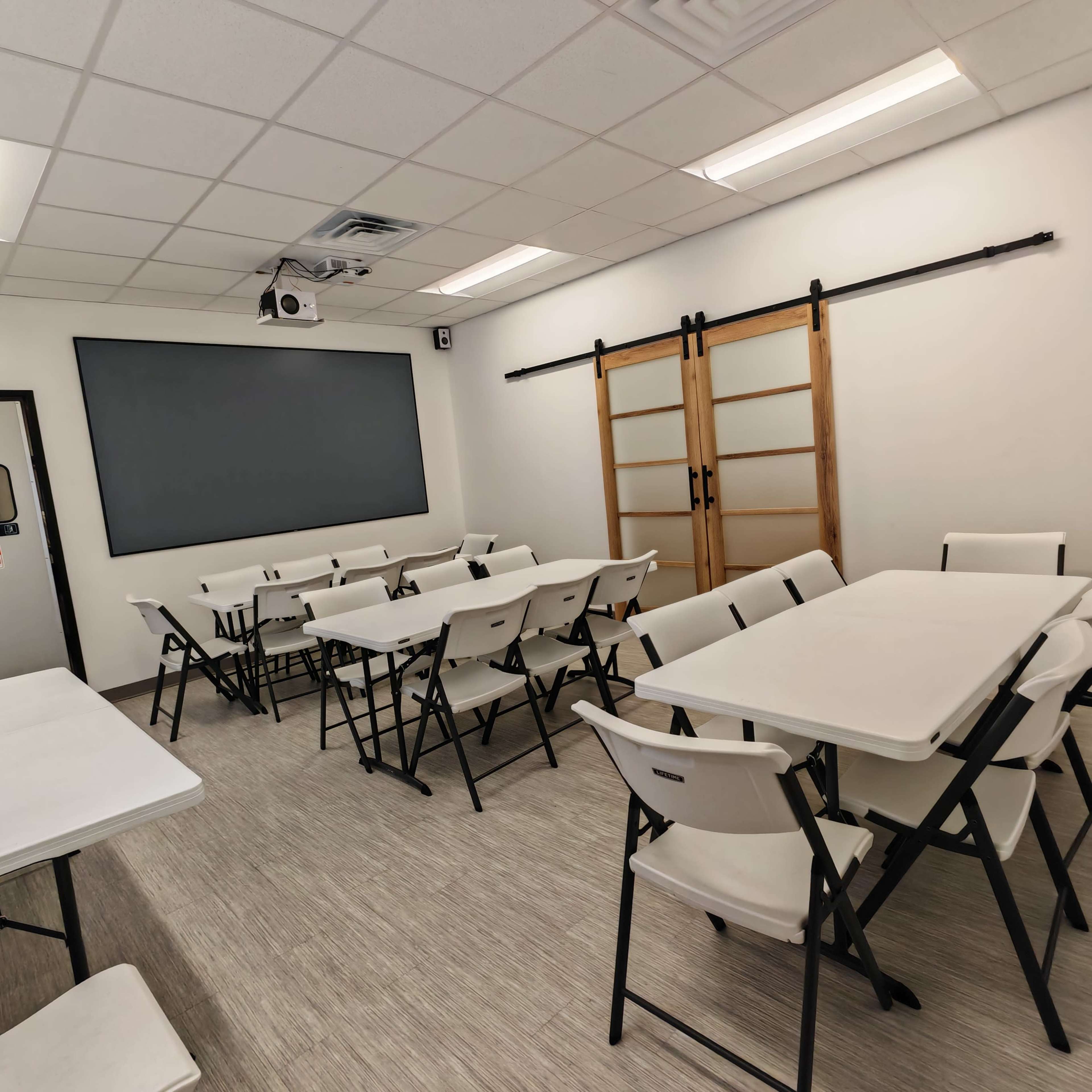 A classroom is arranged with several folding tables and chairs, featuring a projector screen and sliding glass doors.