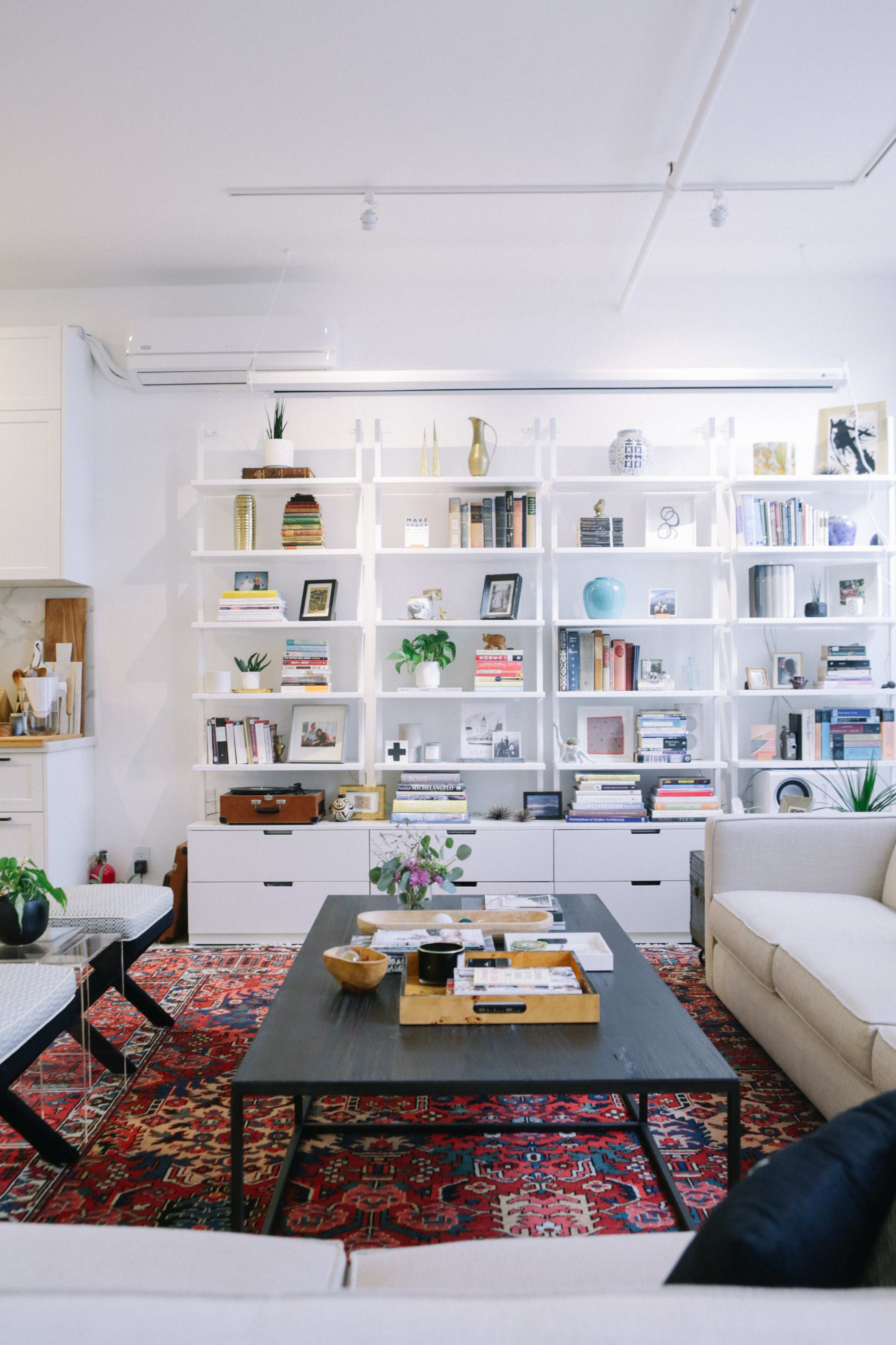 The living room features a spacious bookshelf filled with books and decorative items, a coffee table at the center, and a patterned rug on the floor.