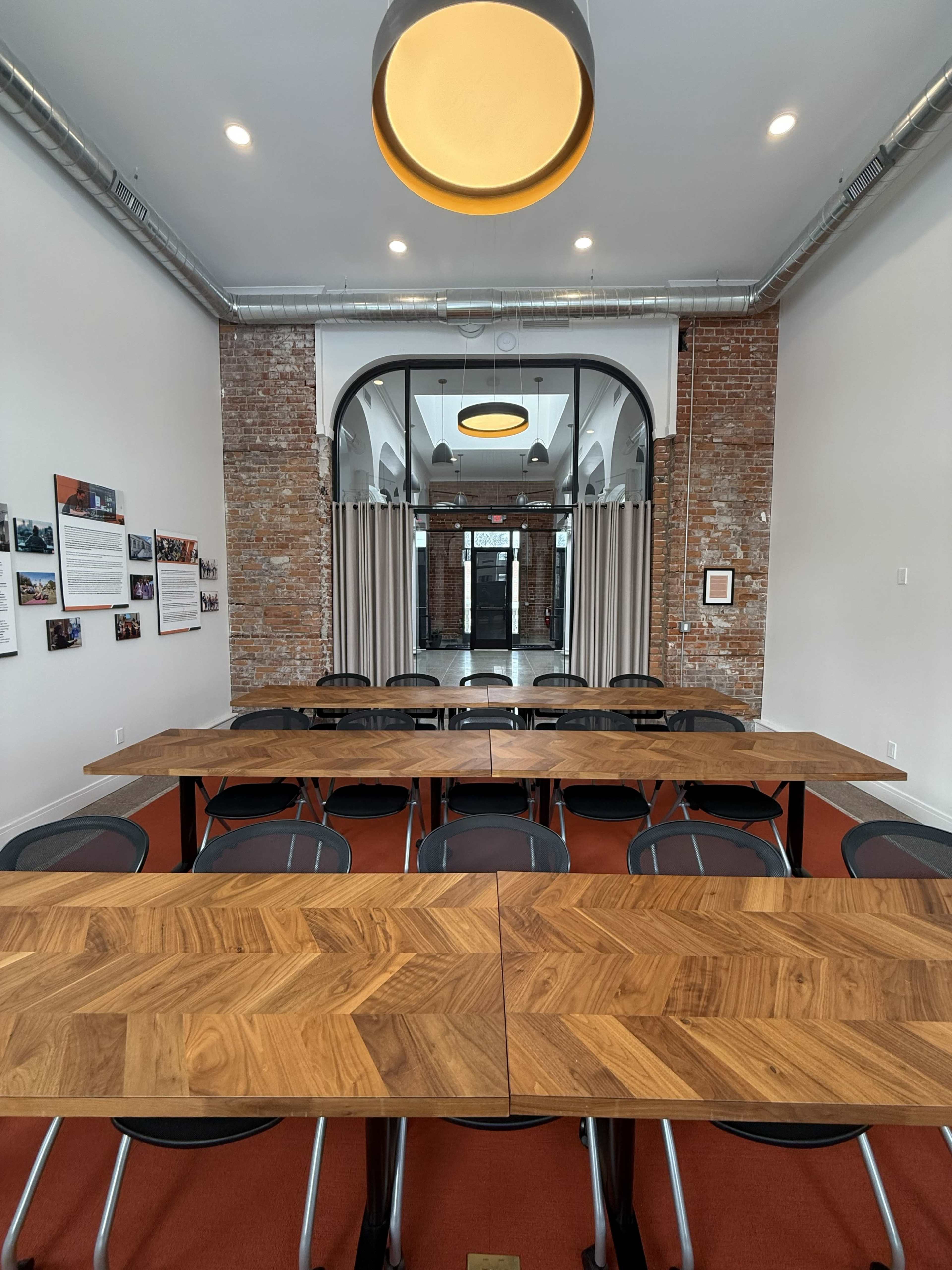 A conference room features multiple wooden tables arranged in a U-shape, with brick walls and large windows providing natural light.