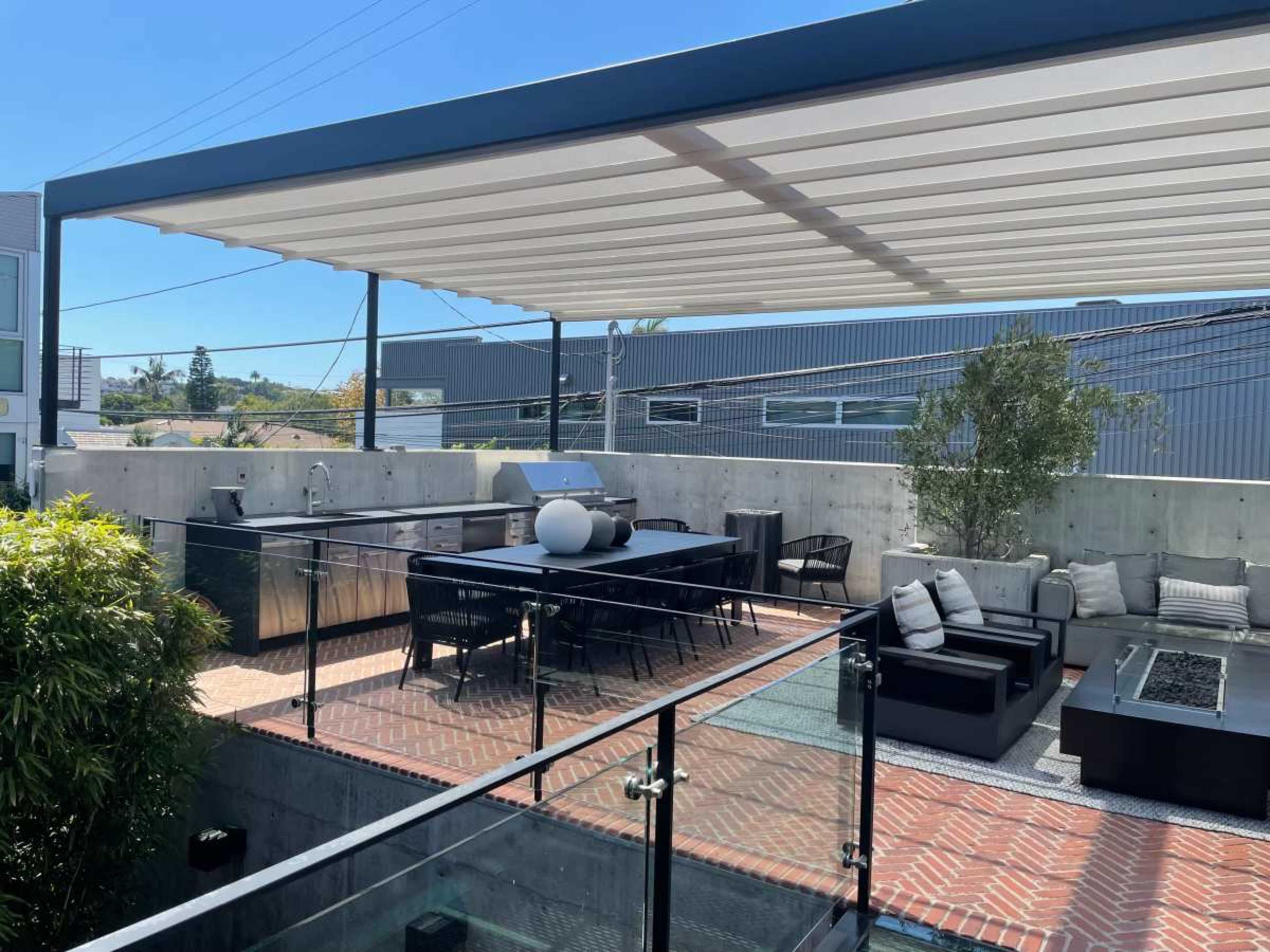 A rooftop patio with a covered seating area, an outdoor kitchen, and a dining table surrounded by concrete walls and greenery.