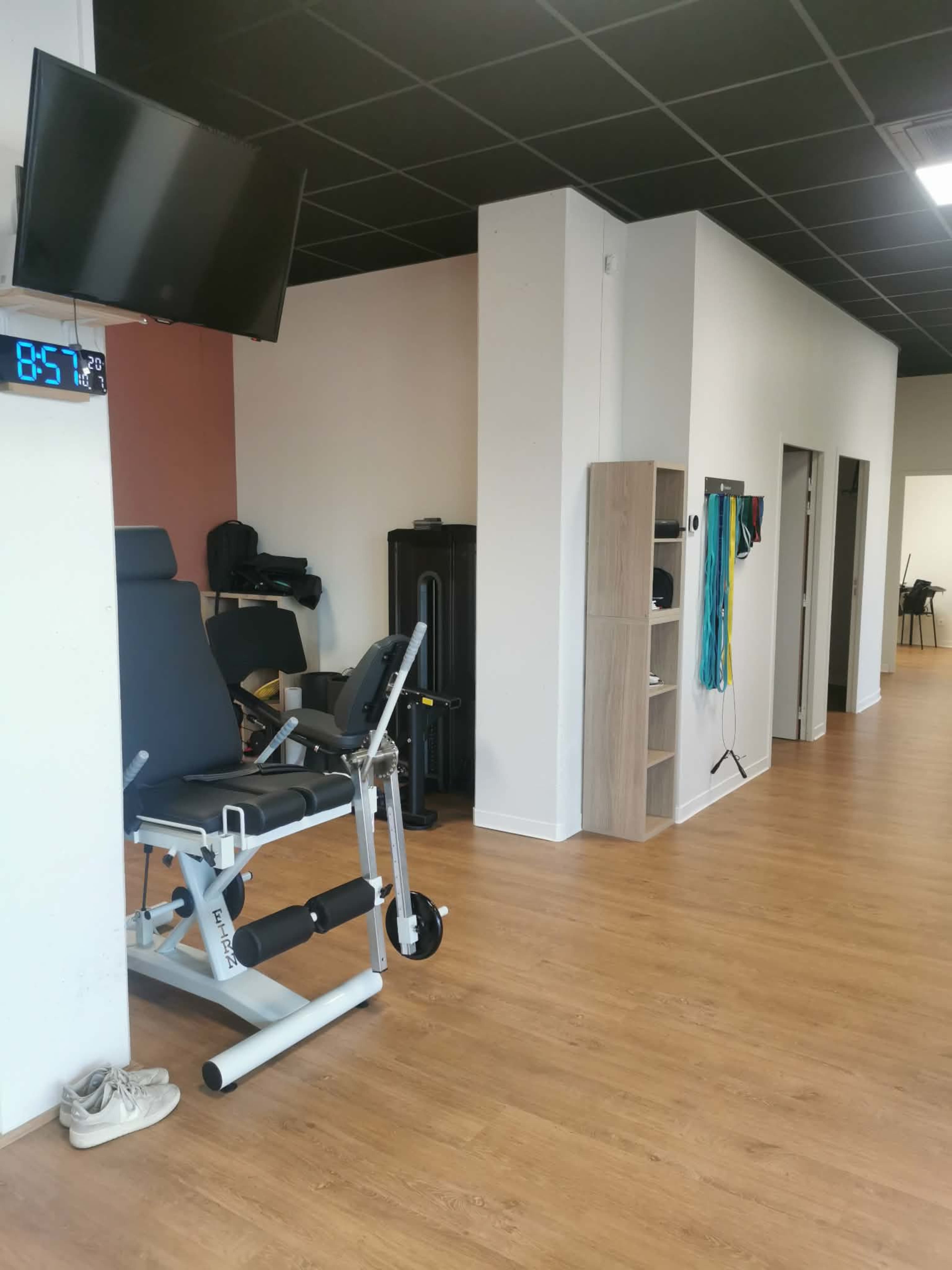 The image shows an exercise area featuring a multifunctional workout machine, a TV mounted on the wall, and shelves with exercise equipment, all set in a spacious, well-lit room with wooden flooring.