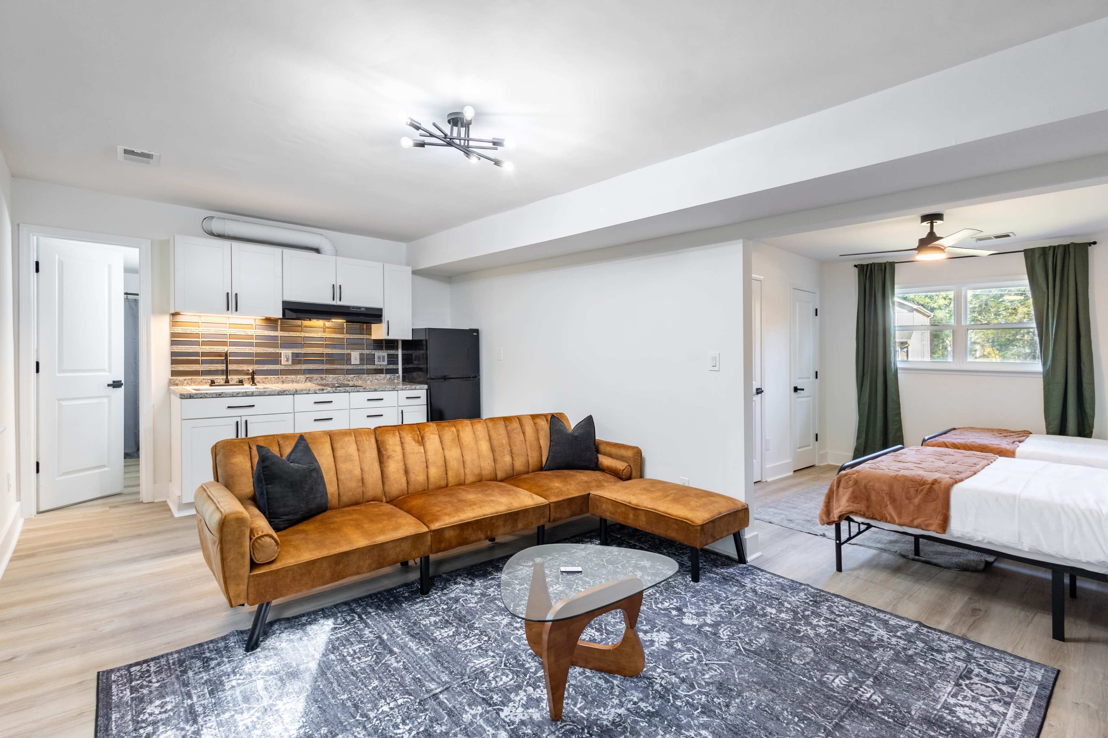 The image shows a modern studio apartment featuring a brown sectional sofa, a glass coffee table, a kitchenette with black appliances, and two separate beds with brown and white linens.
