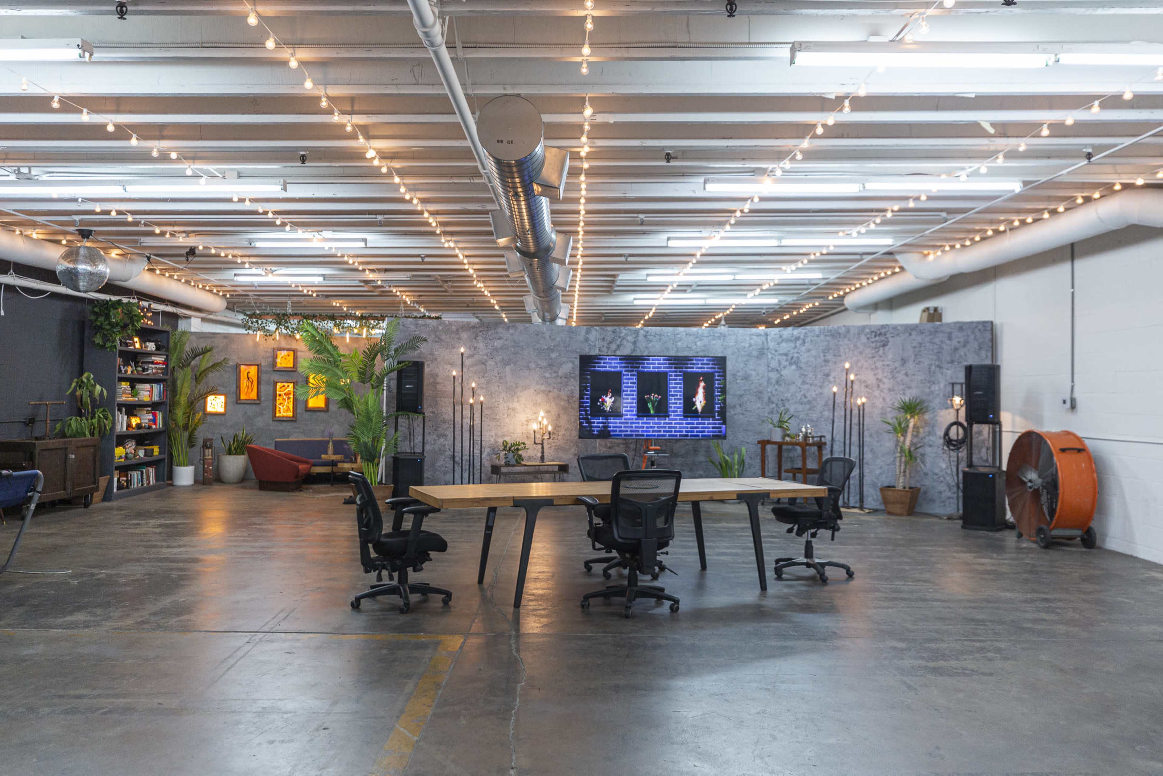 A spacious industrial-style room features a long wooden table surrounded by office chairs, with plants and a large screen displaying content against a textured backdrop.