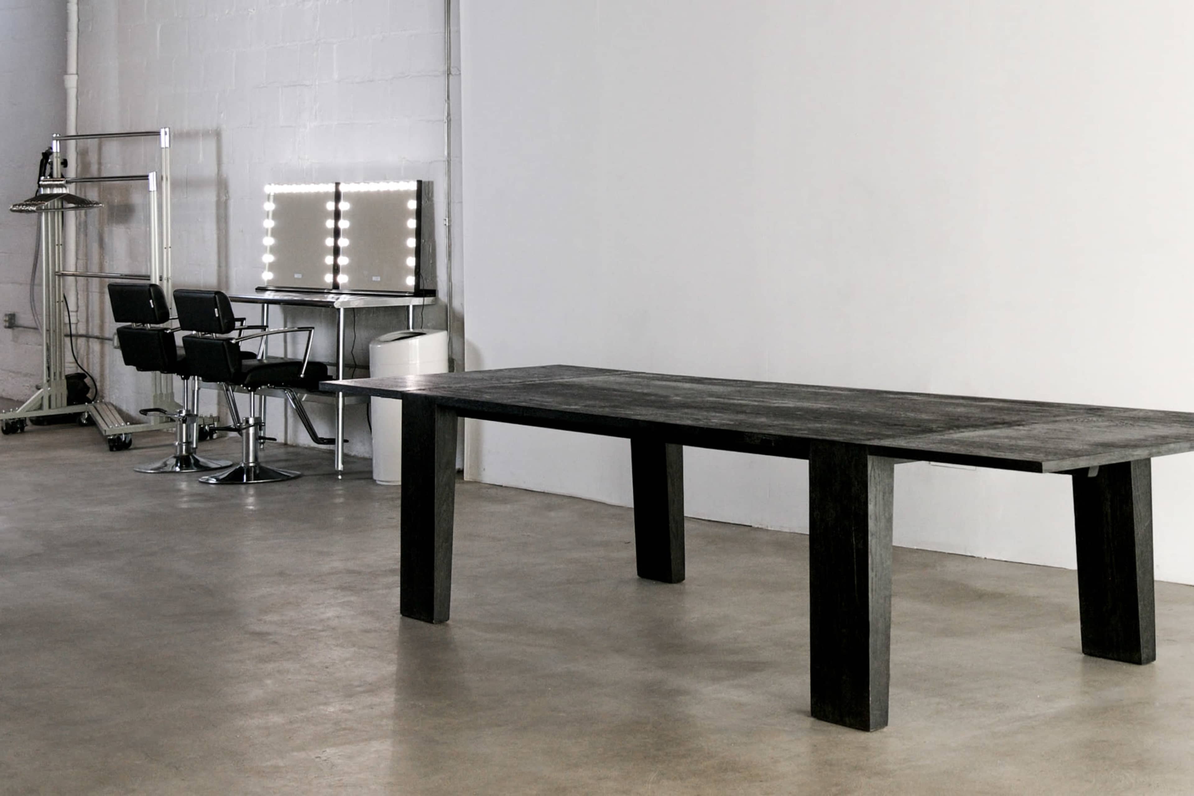 The image features a large black wooden table in an industrial space with a minimalist design, accompanied by two black styling chairs and a lighted makeup mirror against a white wall.