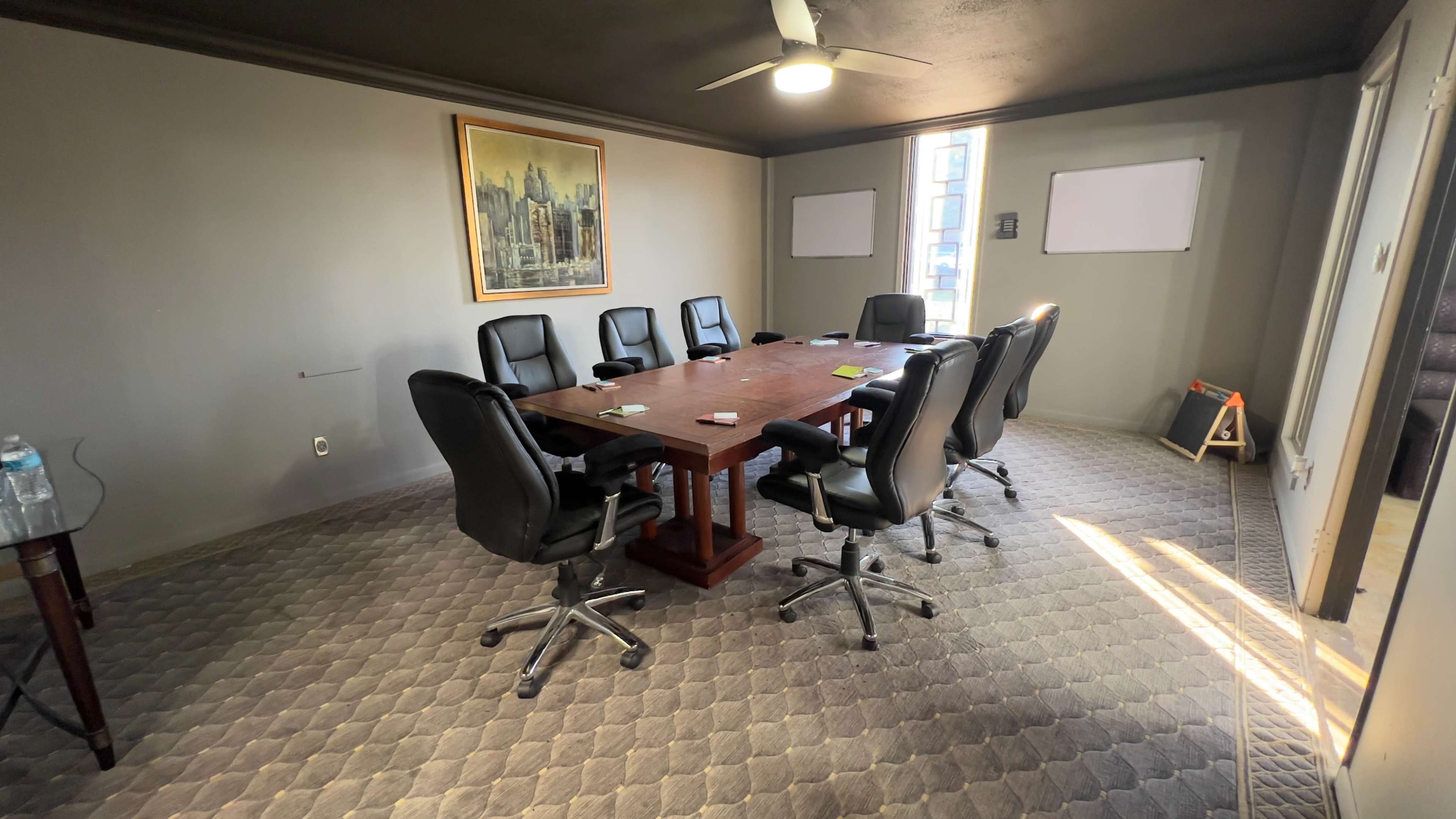 A conference room with a large wooden table surrounded by eight black leather chairs, featuring a painting on the wall and two whiteboards.