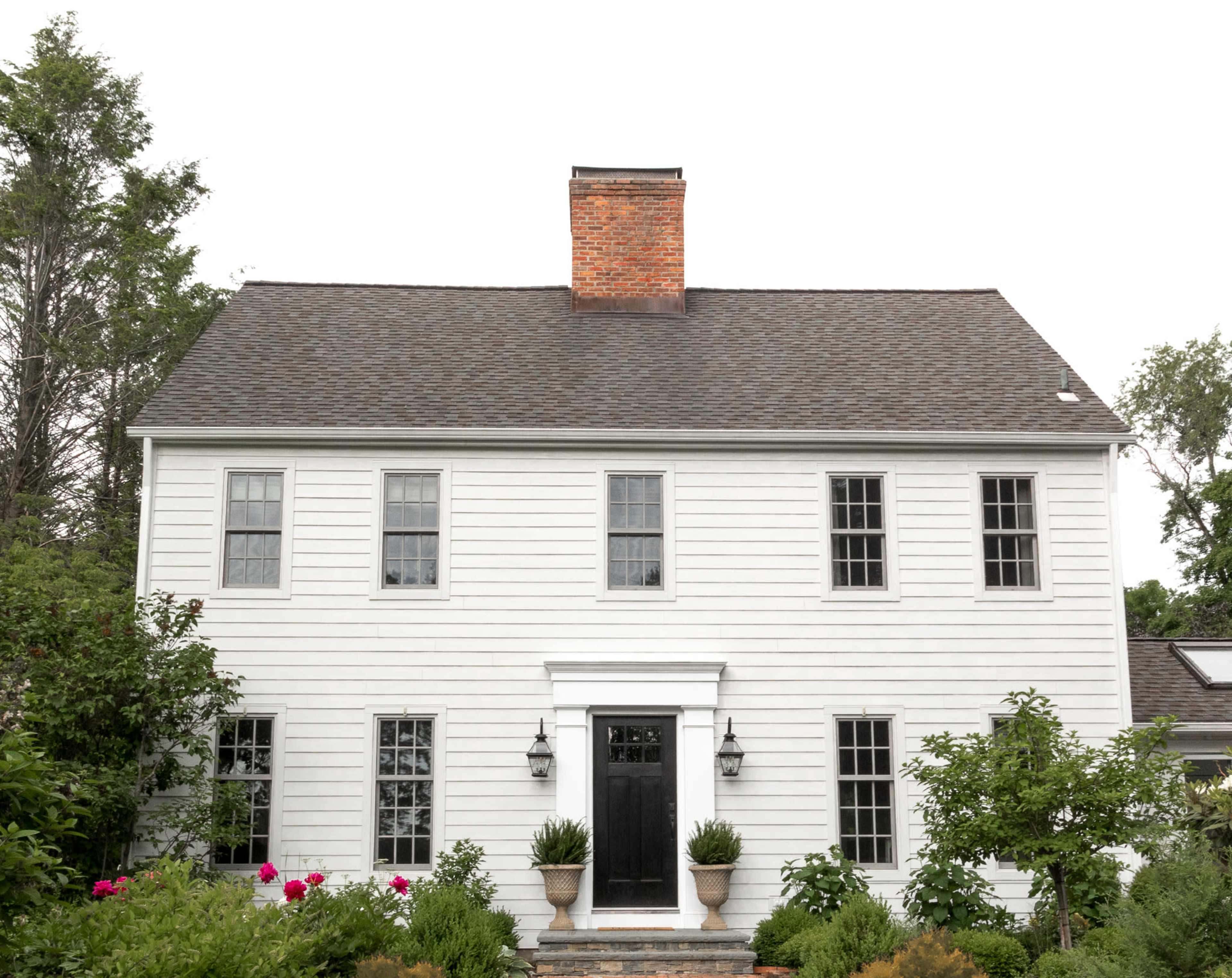 A white wooden house with a central dark door and large windows is surrounded by manicured plants and flowers.