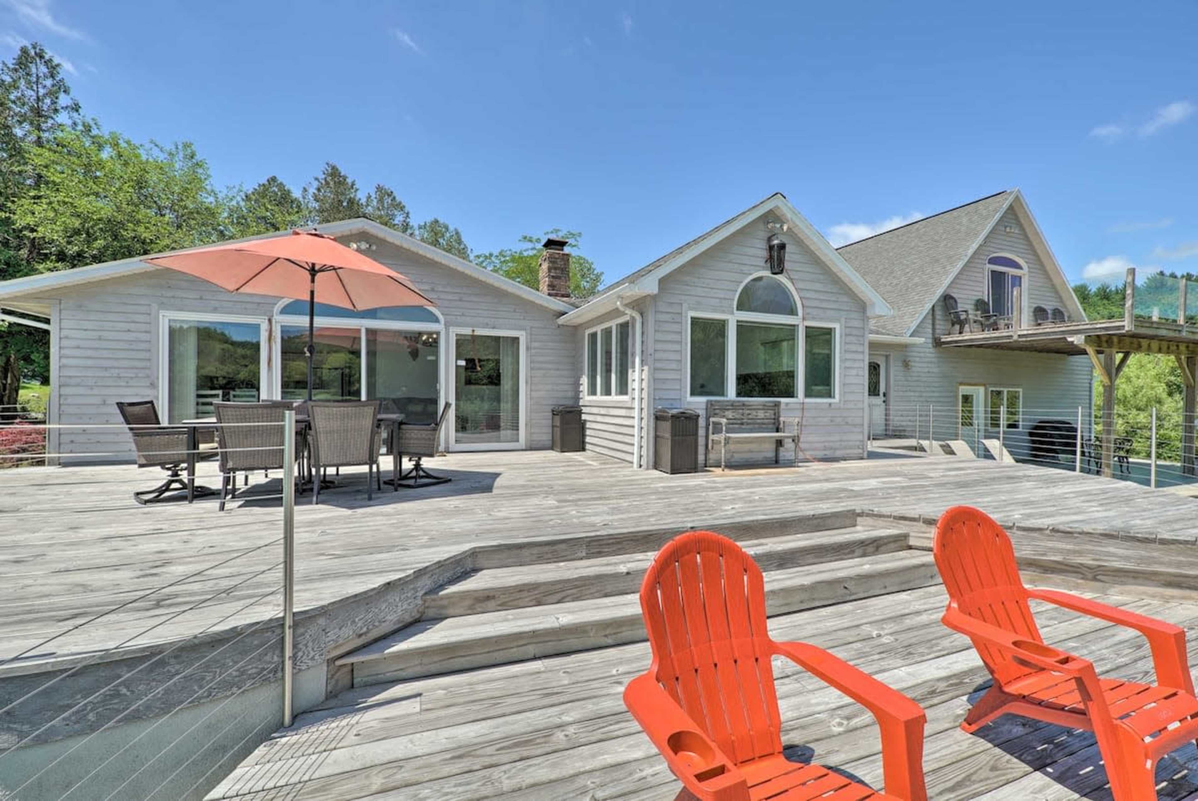 A large wooden deck with two orange chairs and a dining area, adjacent to a modern house surrounded by trees and clear blue skies.