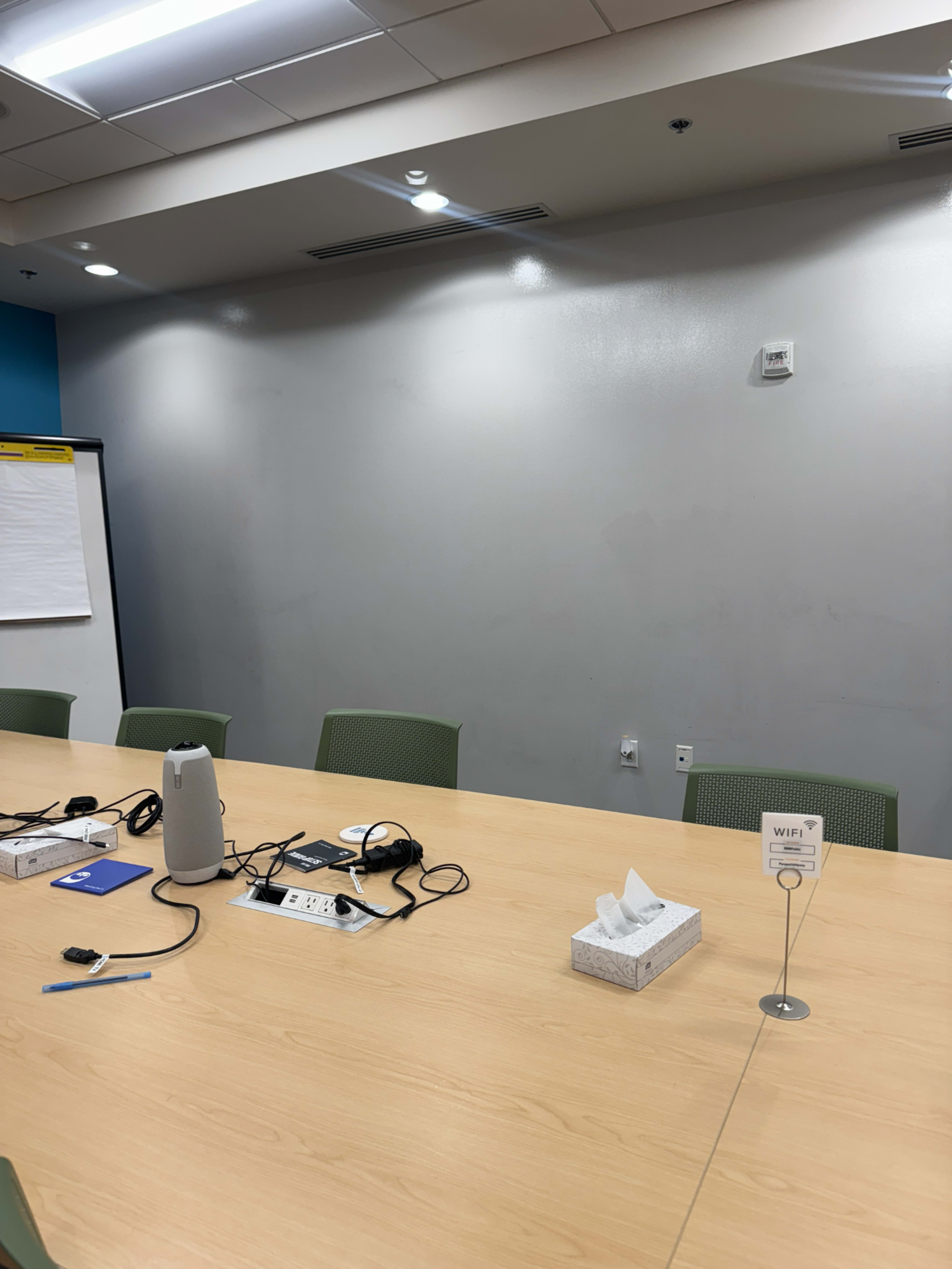 The image shows a conference room with a large wooden table, several chairs, and a whiteboard, along with various electronic devices and a tissue box on the table.