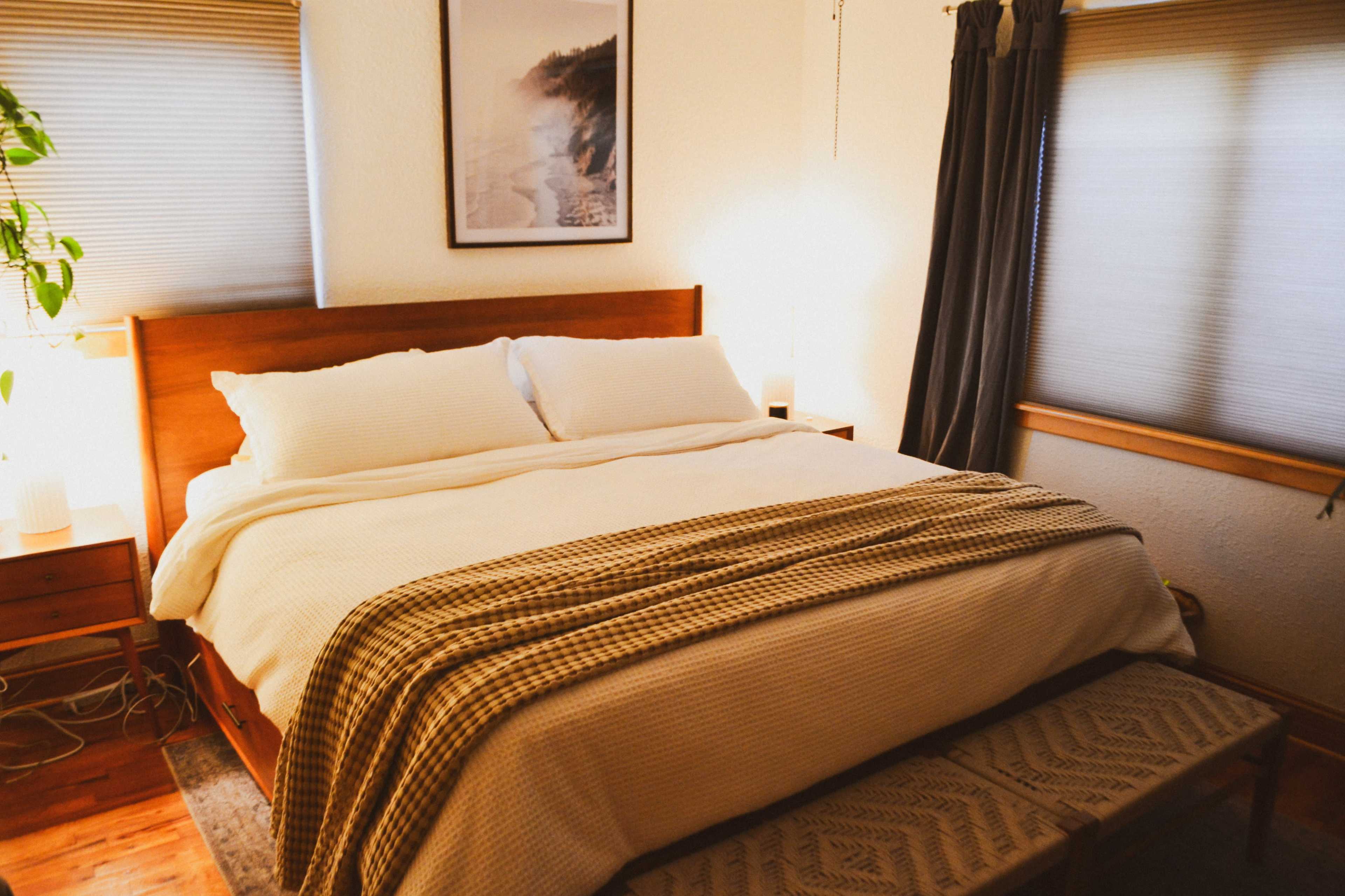 A neatly made bed with white linens is positioned in a room featuring a wooden nightstand, a bench at the foot, and two windows covered with blinds.