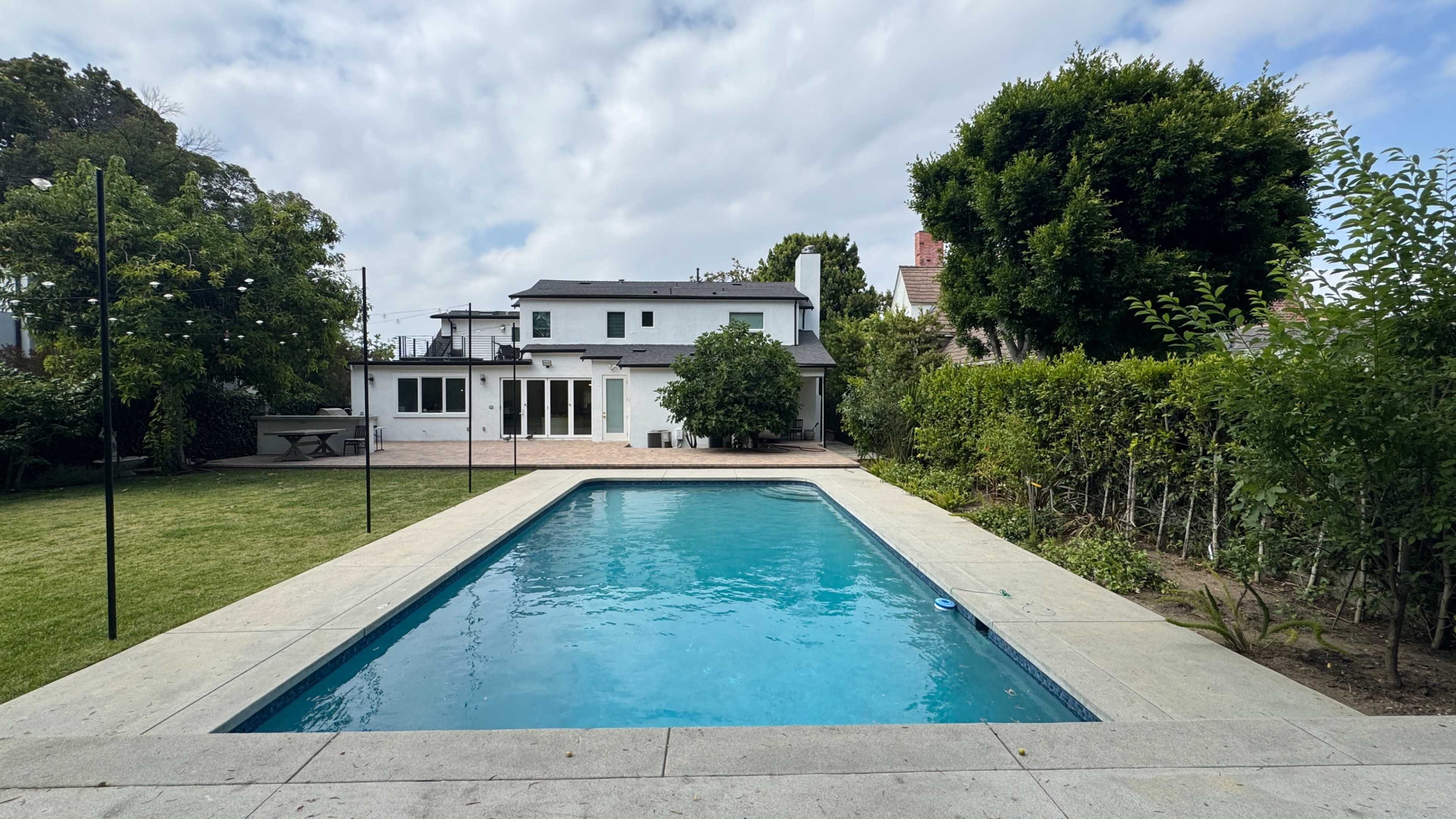 The image shows a backyard with a swimming pool, a two-story house in the background, and greenery surrounding the area.