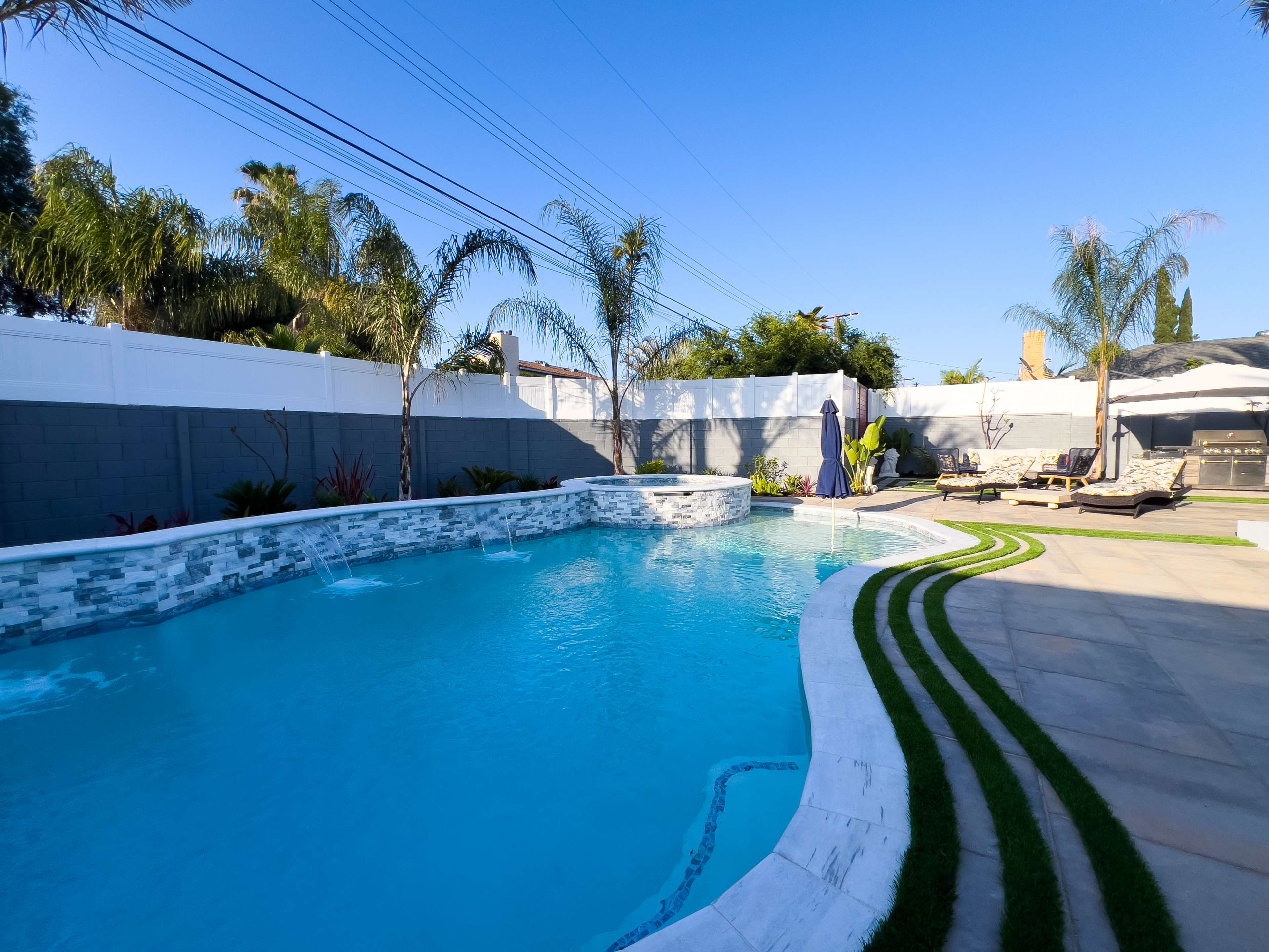 A residential backyard featuring a swimming pool, surrounded by palm trees and lounge chairs, with a well-maintained lawn and stone pathways.