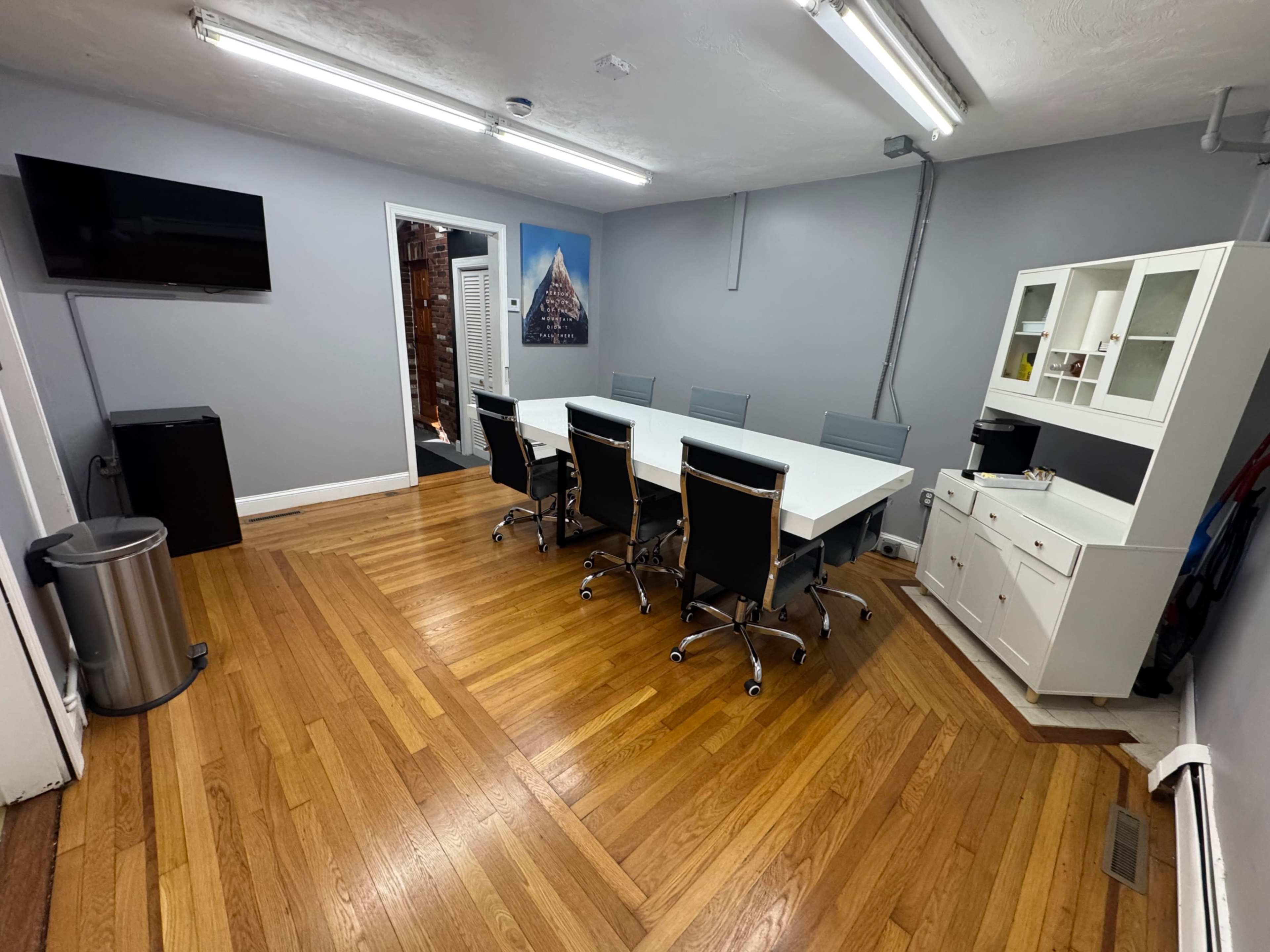 The image shows a modern meeting room with a long white table, six black chairs, a TV on the wall, and a white cabinet in the corner.