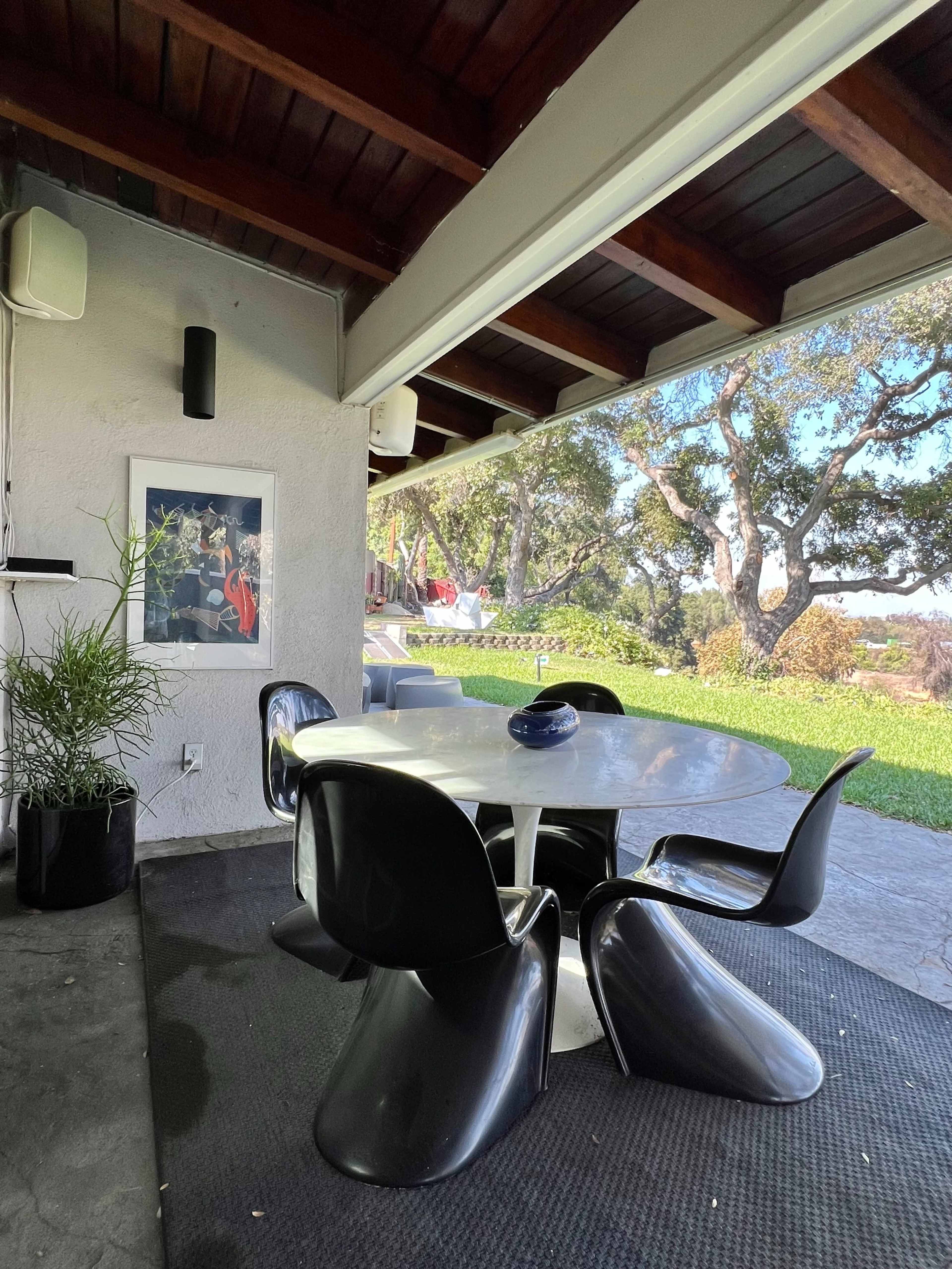 A round table with black chairs is set under a covered patio, overlooking a grassy area and trees in the background.