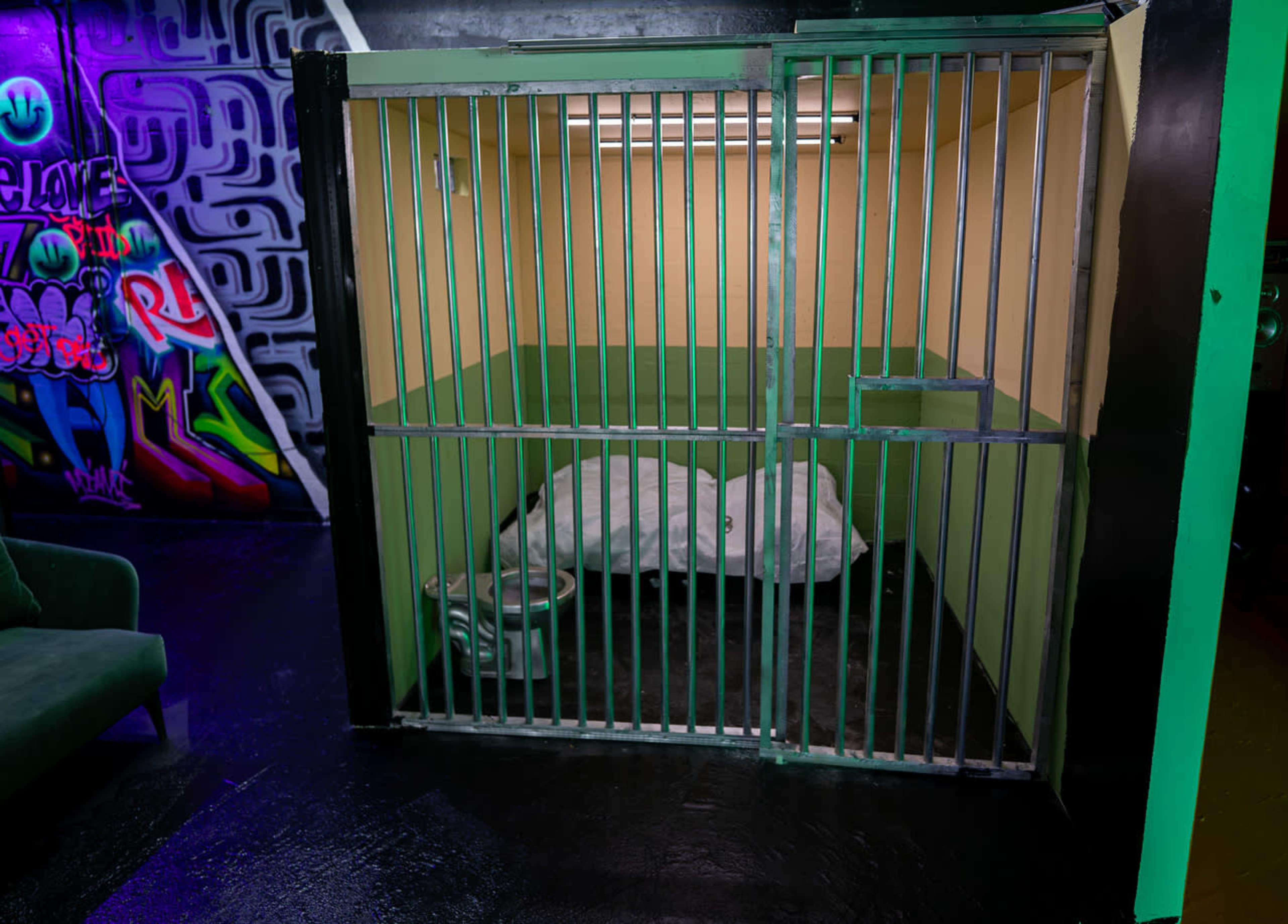 A metal cage with a bed covered by a blanket inside, set against a colorful graffiti wall.