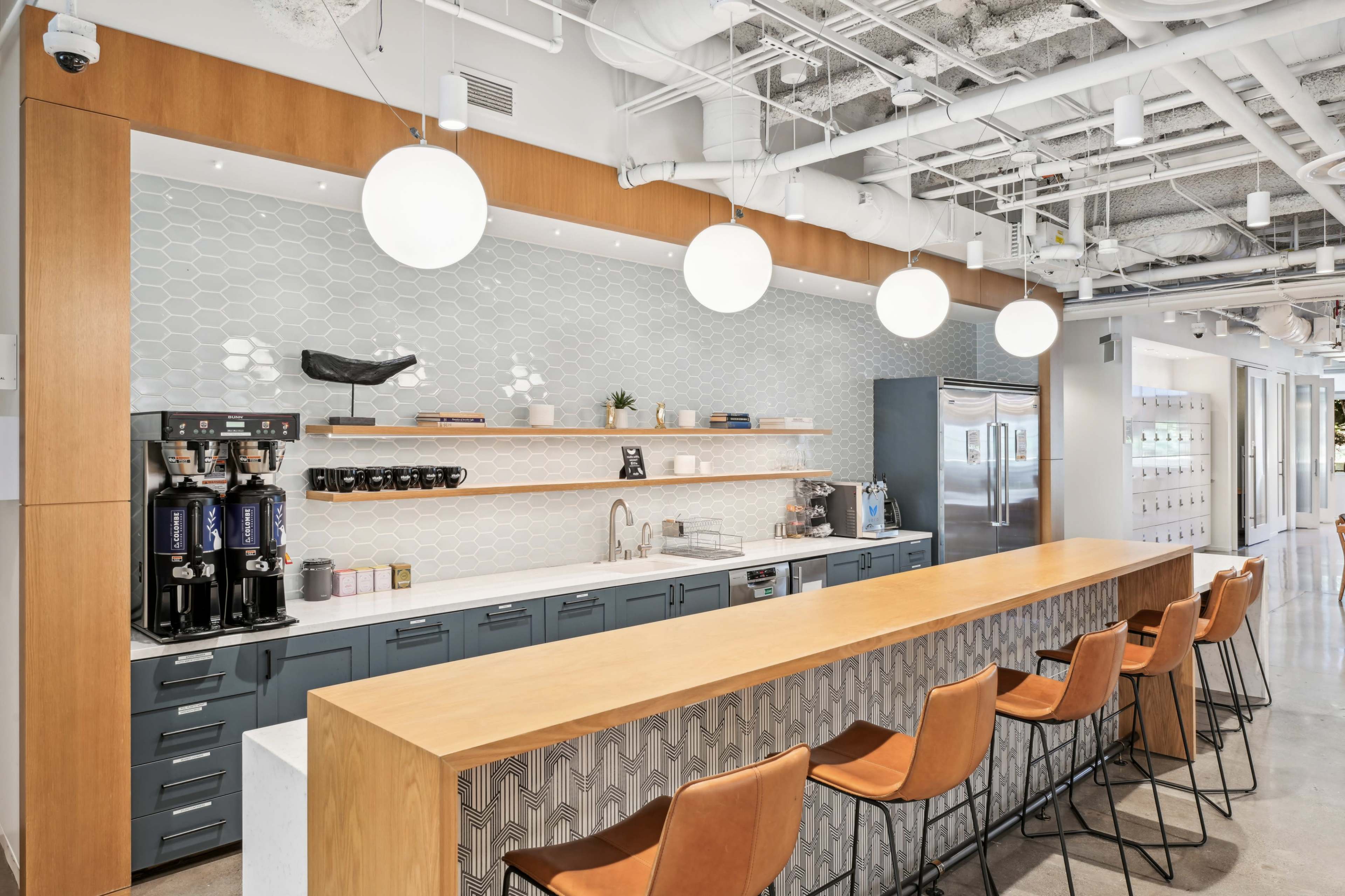A modern kitchen area features a long wooden countertop with barstools, a coffee station, and sleek appliances under circular pendant lights.