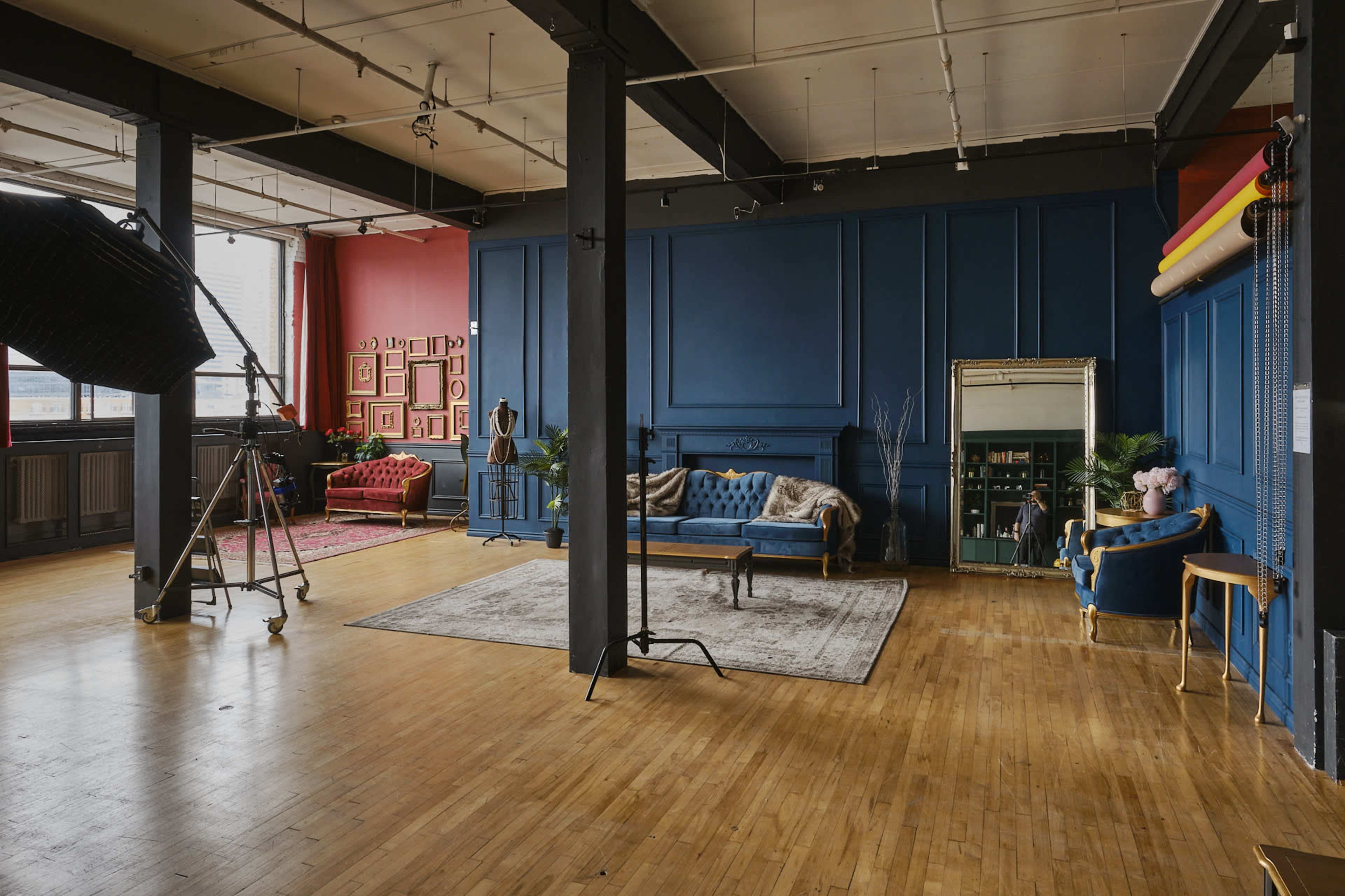 A spacious studio with wooden flooring, featuring a blue and red color scheme, and furnished with elegant sofas, a mirror, and photography equipment.