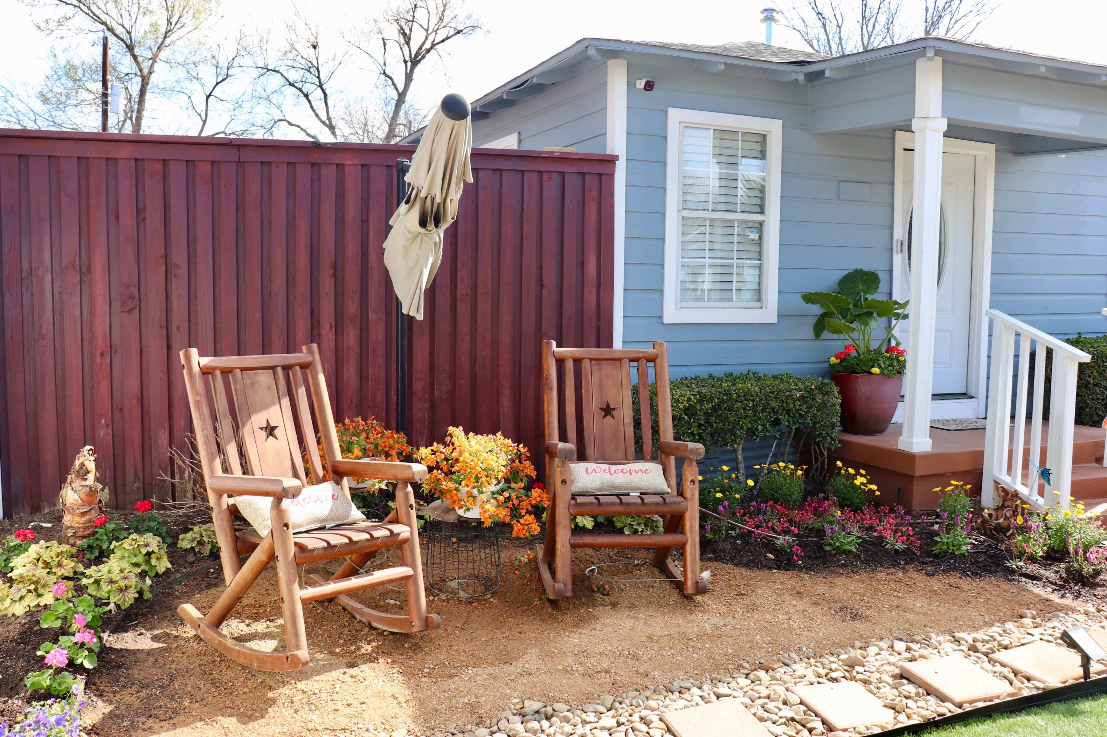 Two rocking chairs with star motifs sit on a gravel path beside a house with blue siding, framed by a wooden fence and colorful flowerbeds.