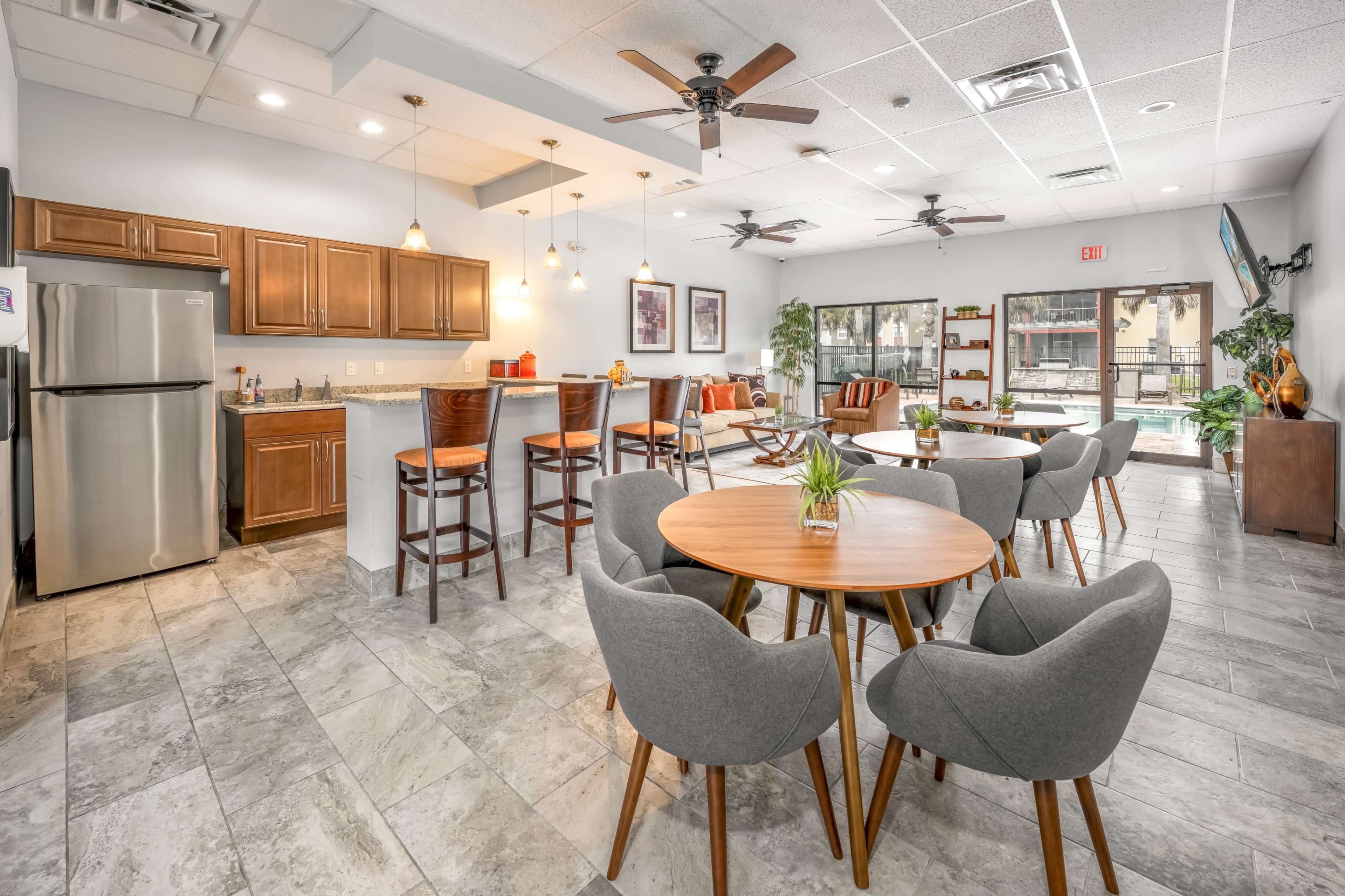 The image shows a modern common area featuring tables with chairs, a kitchenette, and large windows overlooking a pool.