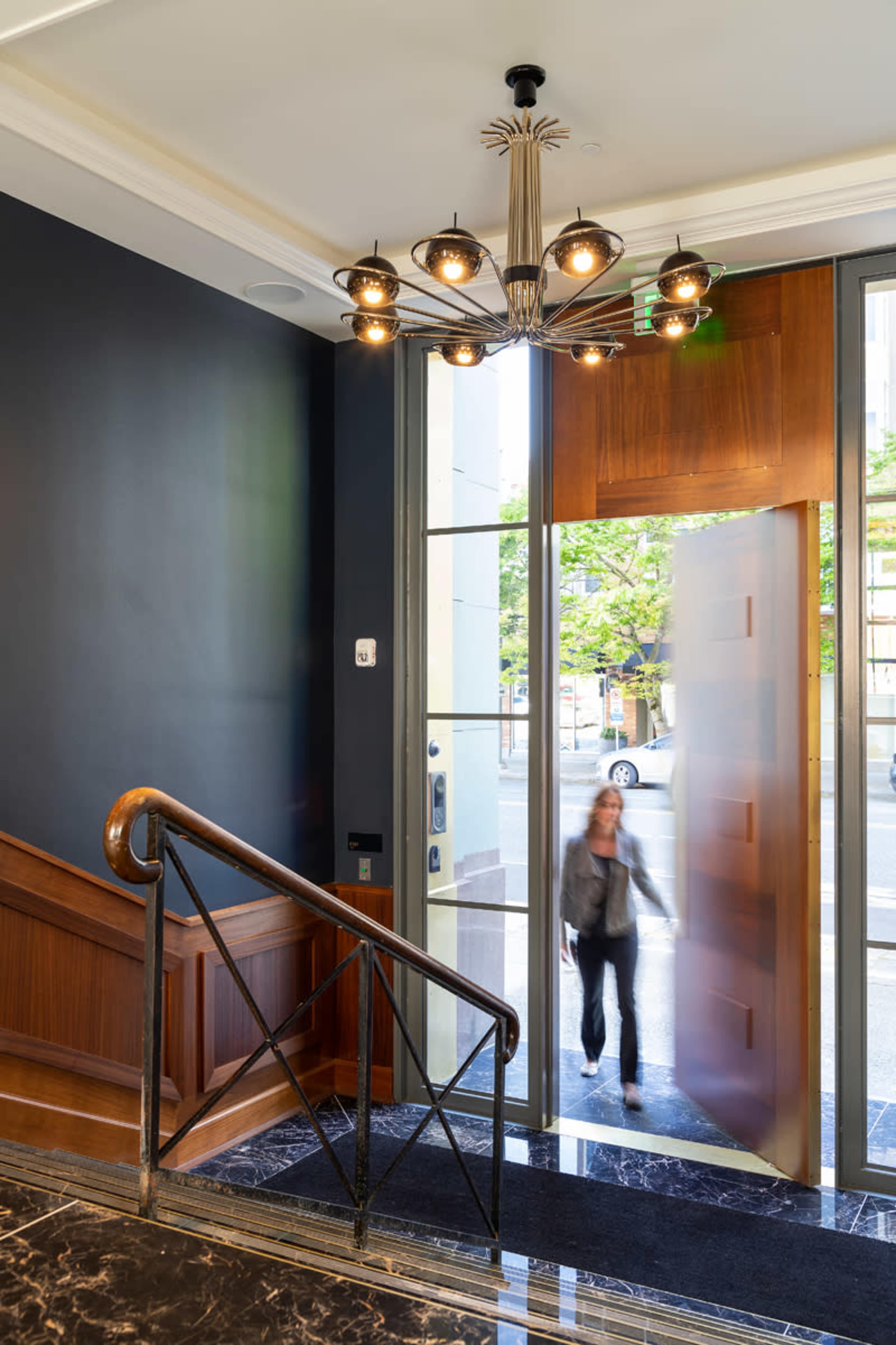 A woman walks through a glass door at the entrance of a building, with a staircase and chandelier visible in the background.