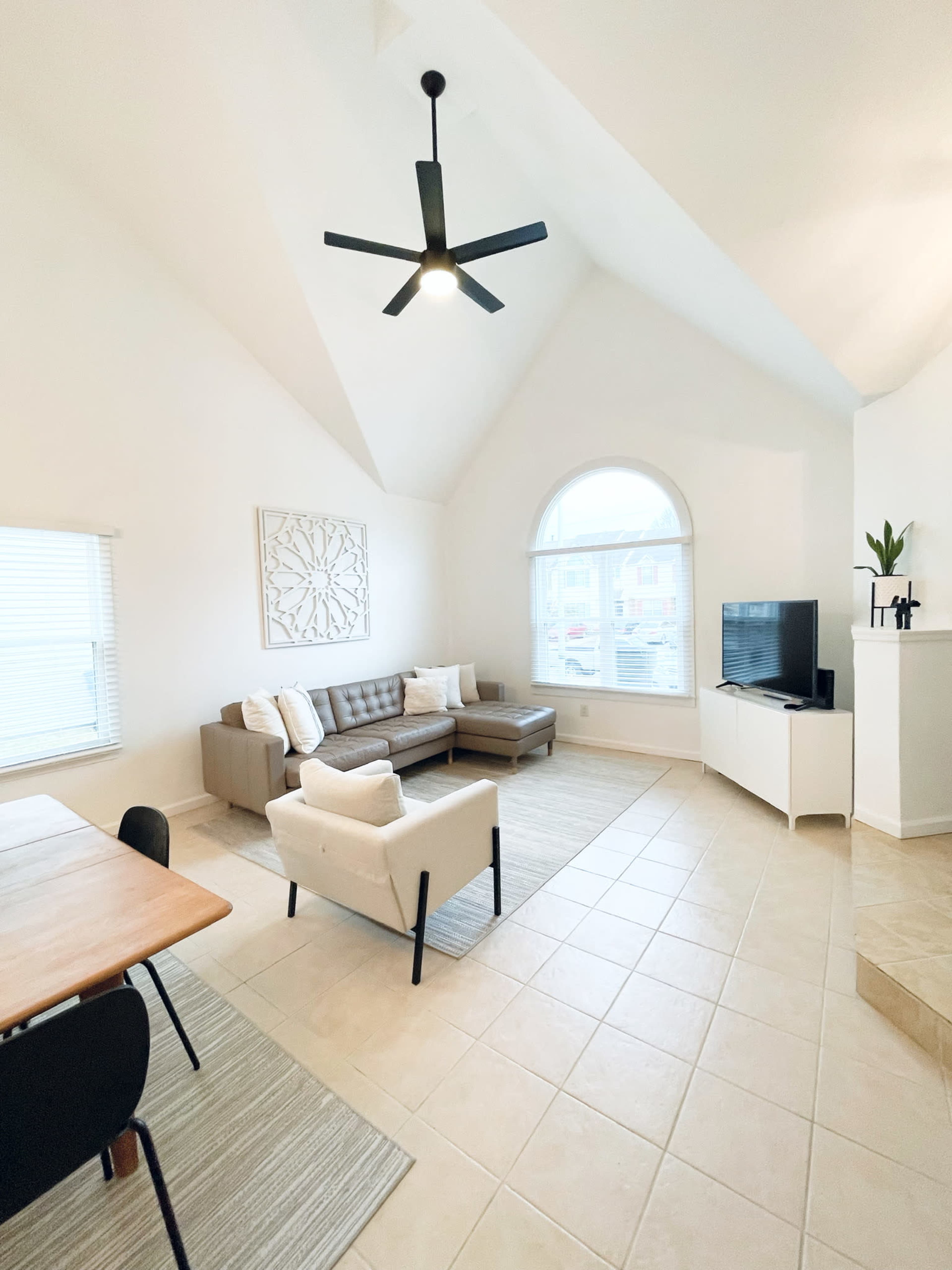 The image shows a bright, modern living room with high ceilings, featuring a sectional sofa, a coffee table, and a television on a white cabinet.