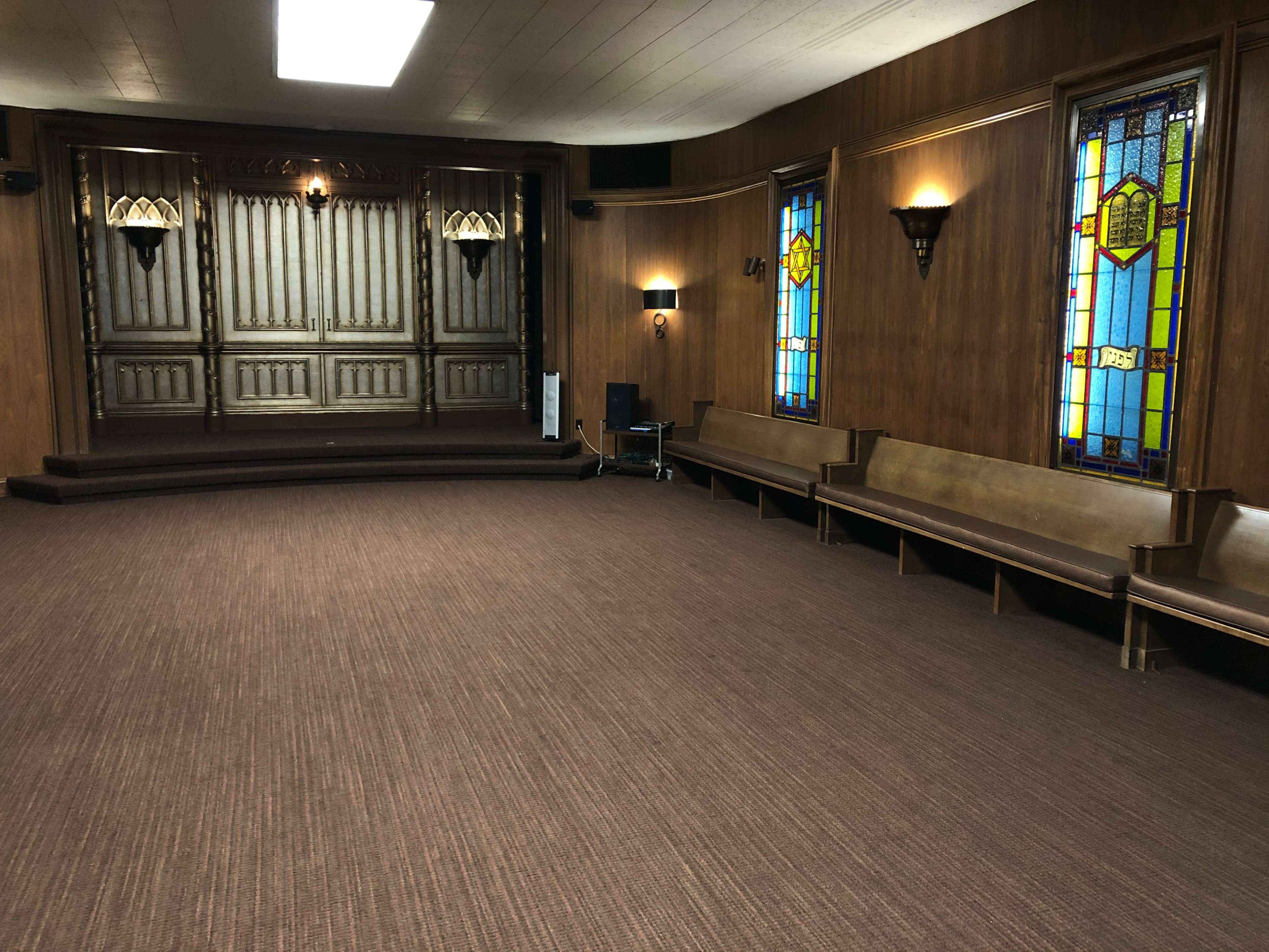 An interior space with wooden walls, benches along the sides, and stained glass windows, featuring a raised stage at the back.