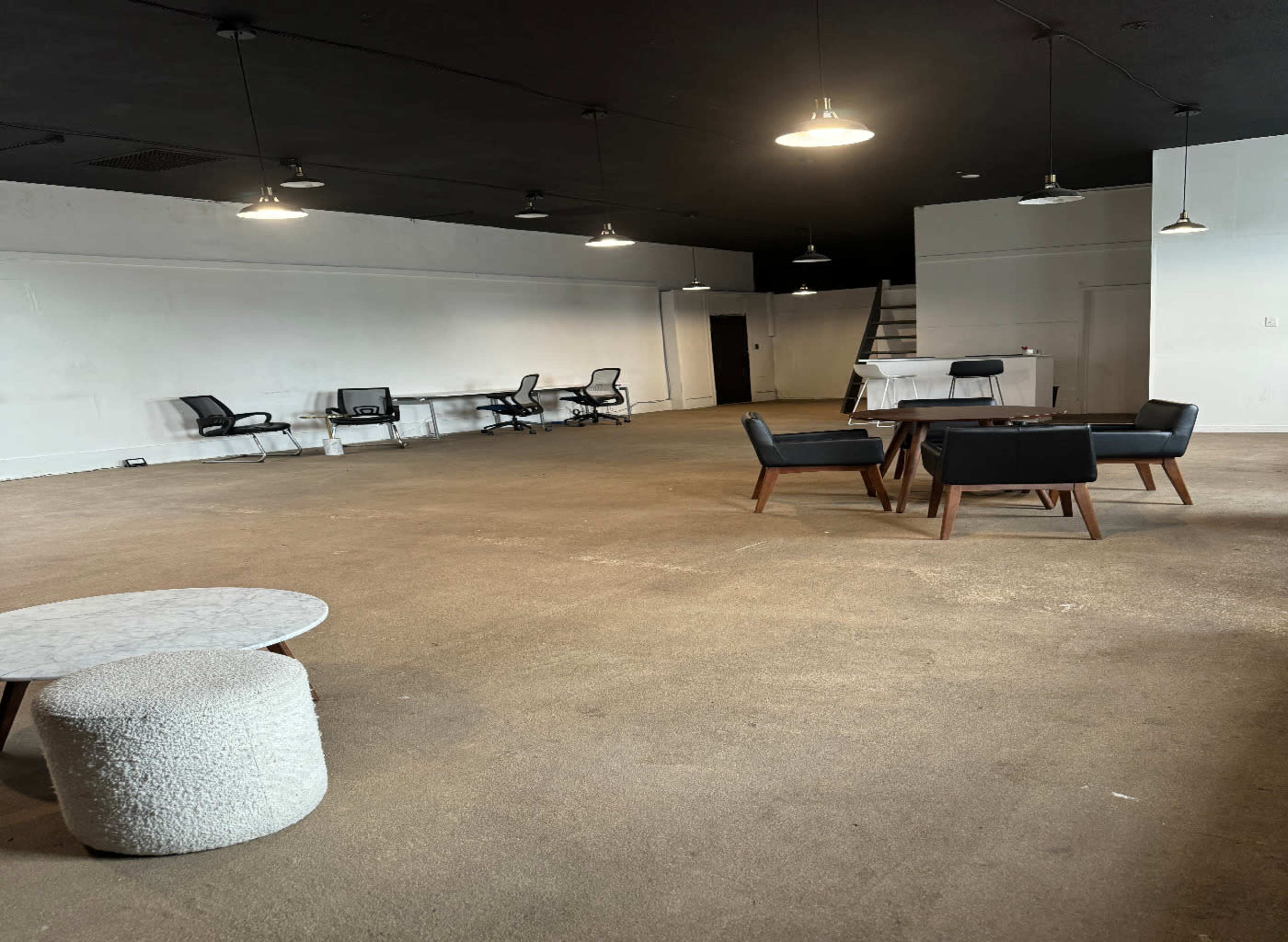 The image shows a spacious, empty room with beige carpet, several chairs, a round table, and a white wall with a staircase in the background.