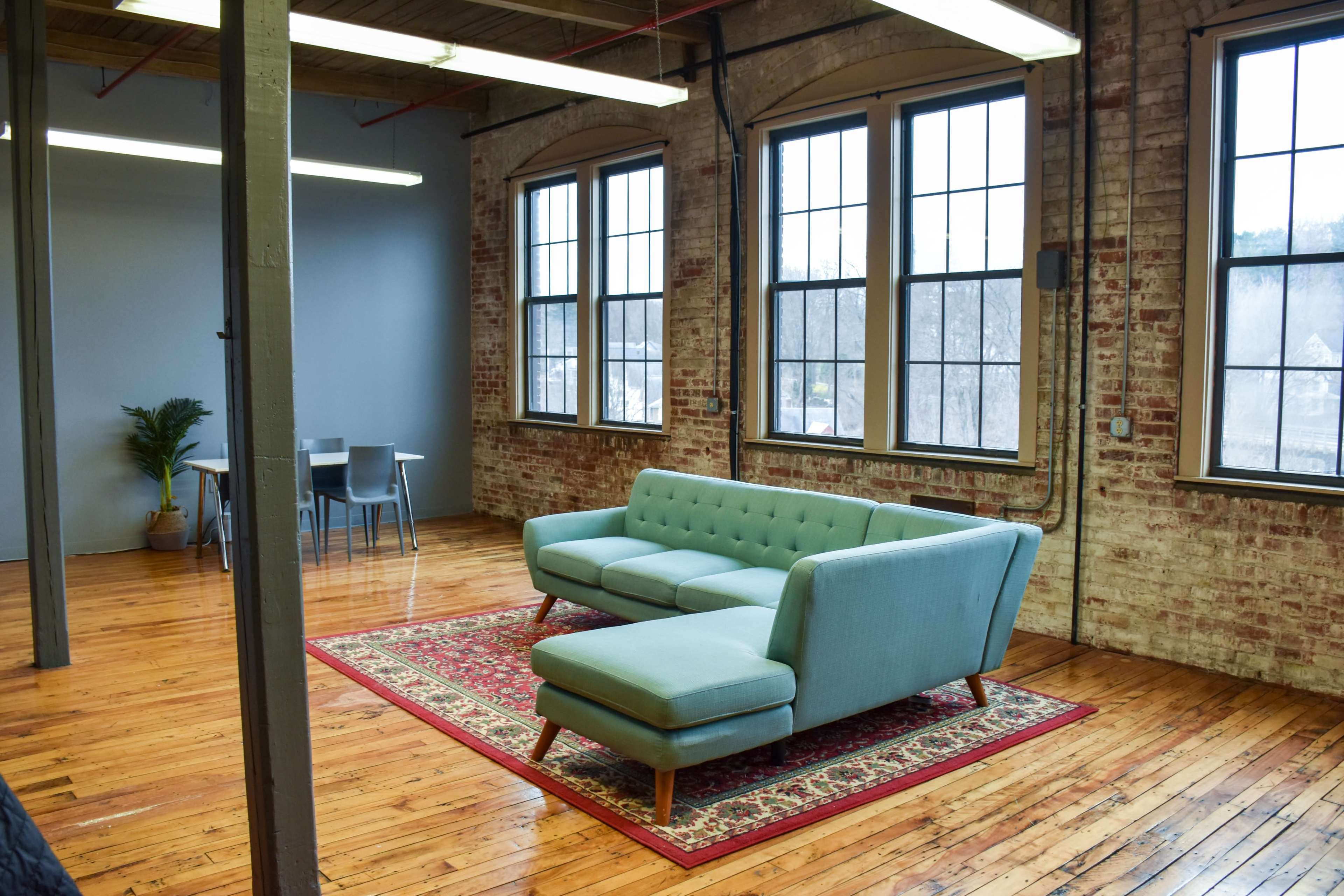 A modern, spacious room with a light blue couch, a patterned rug, large windows, and wooden floors.