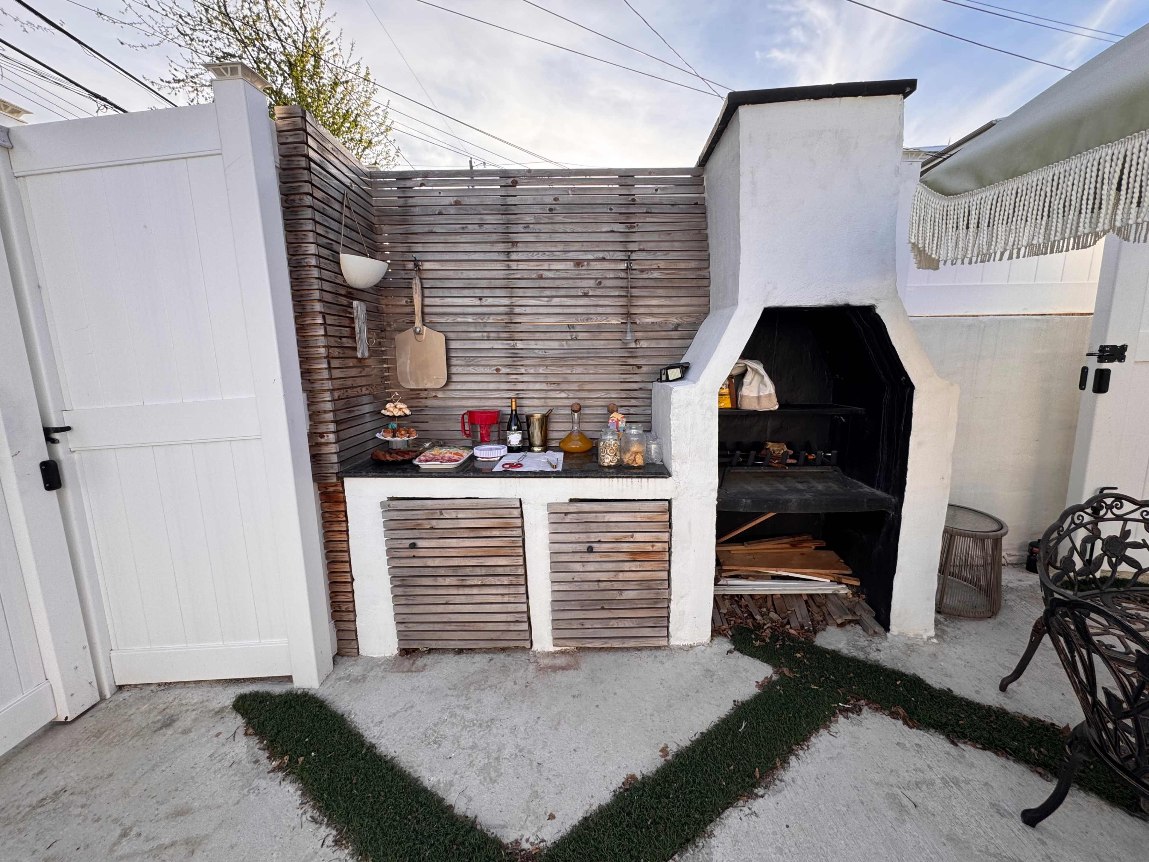 The image features an outdoor kitchen area with a grill, countertop, and wooden storage enclosed by a fence and a white wall.
