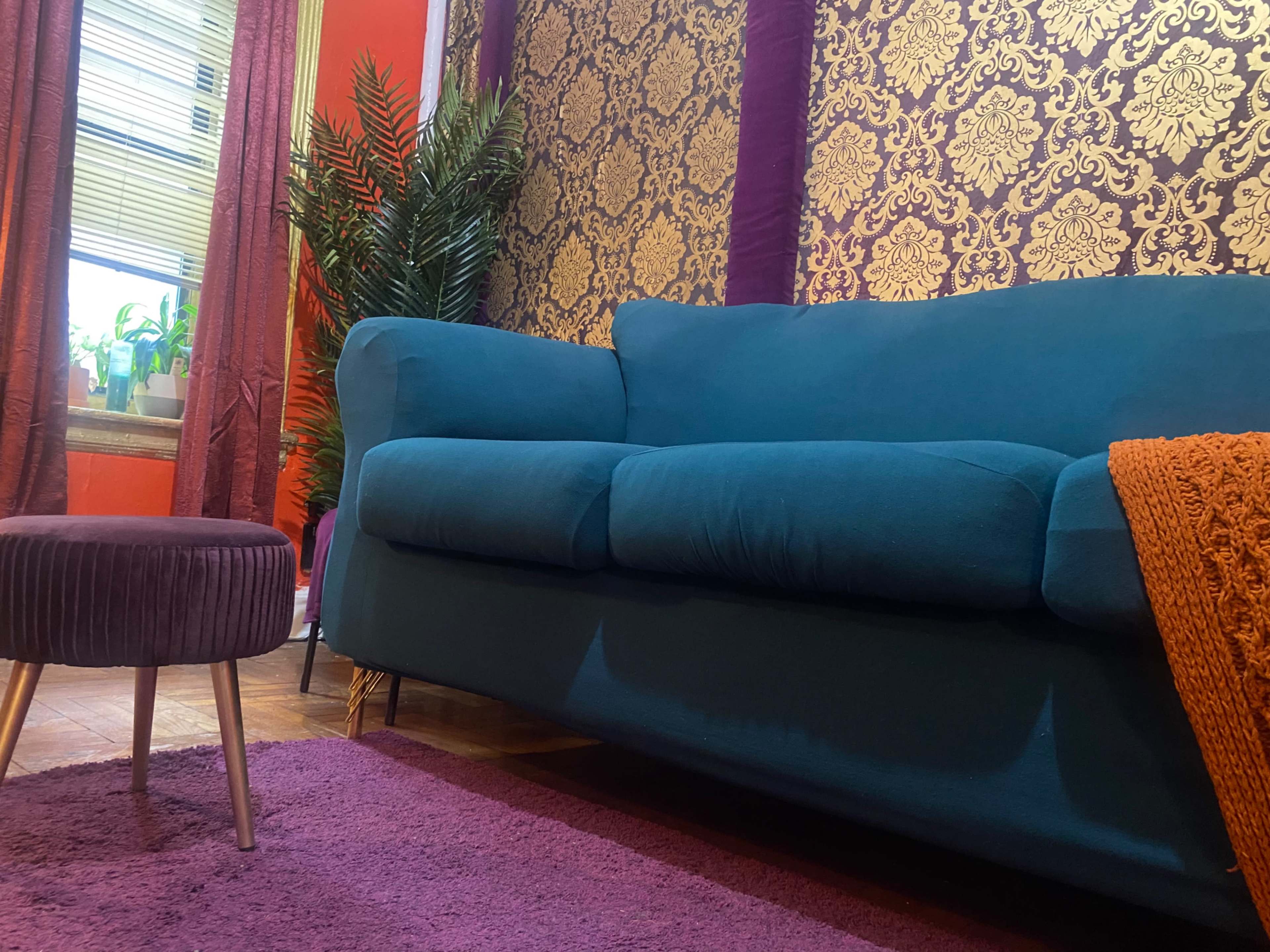A teal couch with a round, pleated ottoman on a purple rug, set against a backdrop of ornate gold wallpaper and a potted plant by the window.