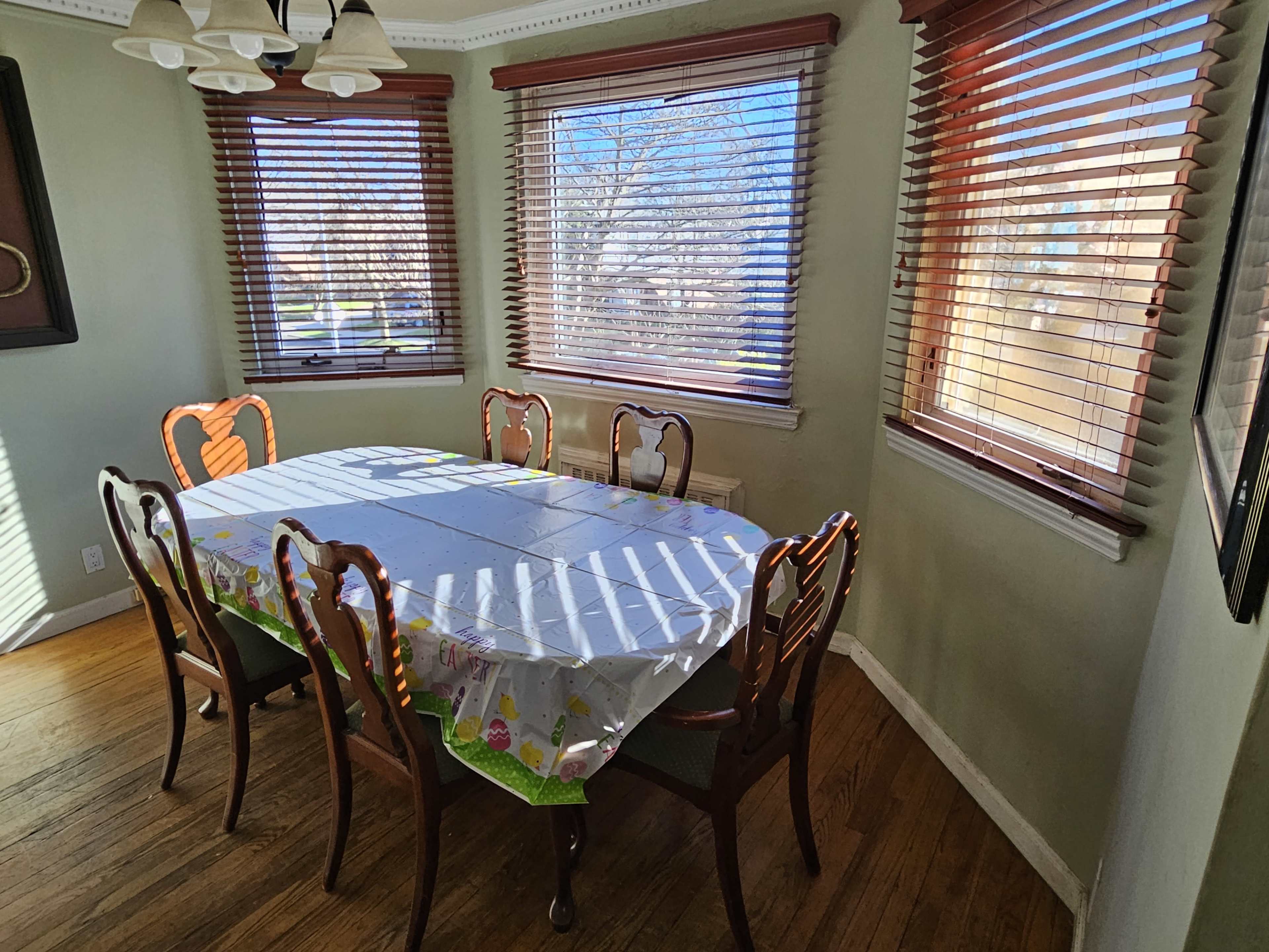 A dining table with six chairs is set in a well-lit room featuring window blinds and hardwood flooring.