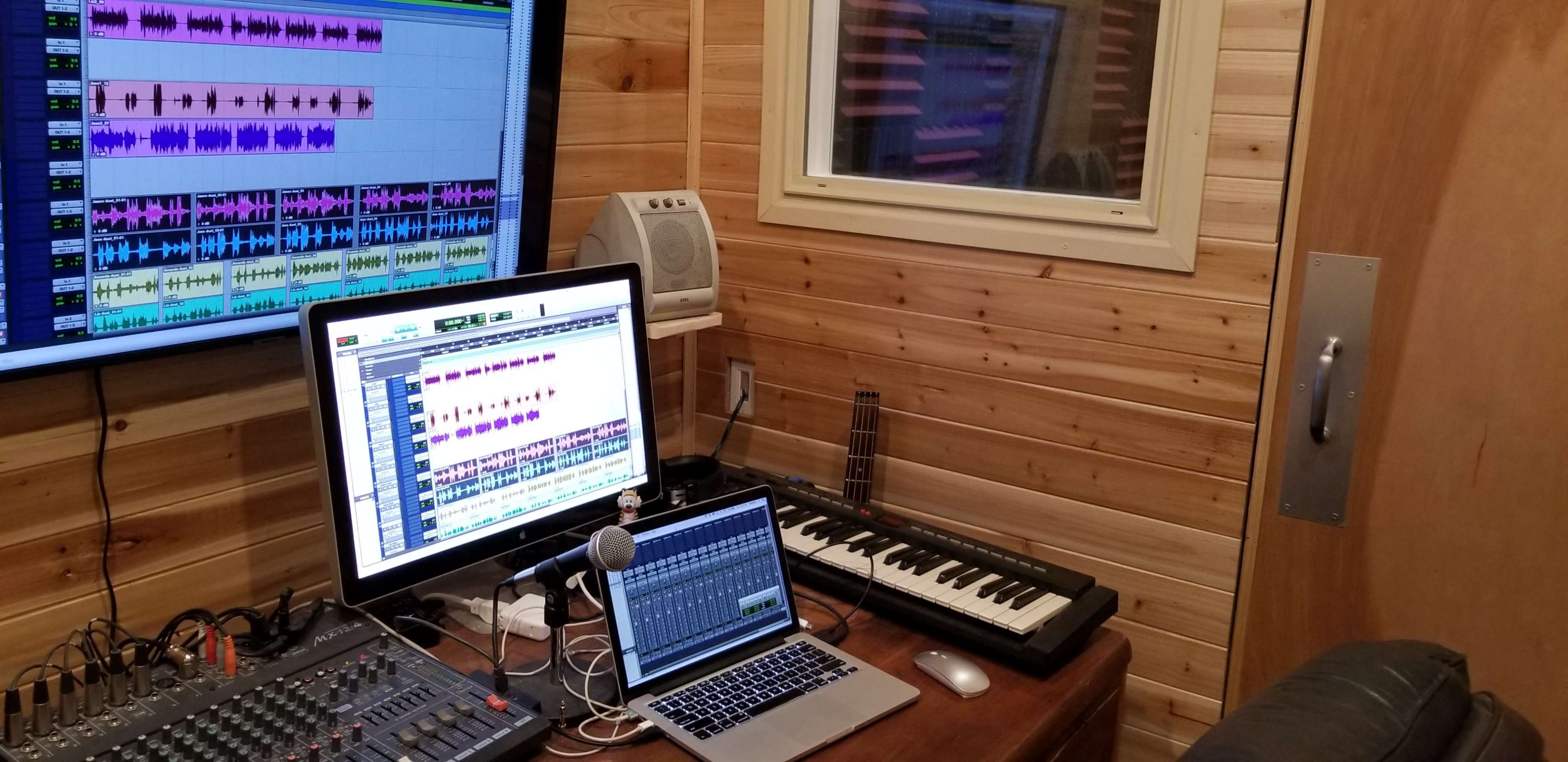 A music production studio with a large monitor displaying audio tracks, a smaller laptop, a mixing console, a keyboard, and a microphone on a wooden desk.