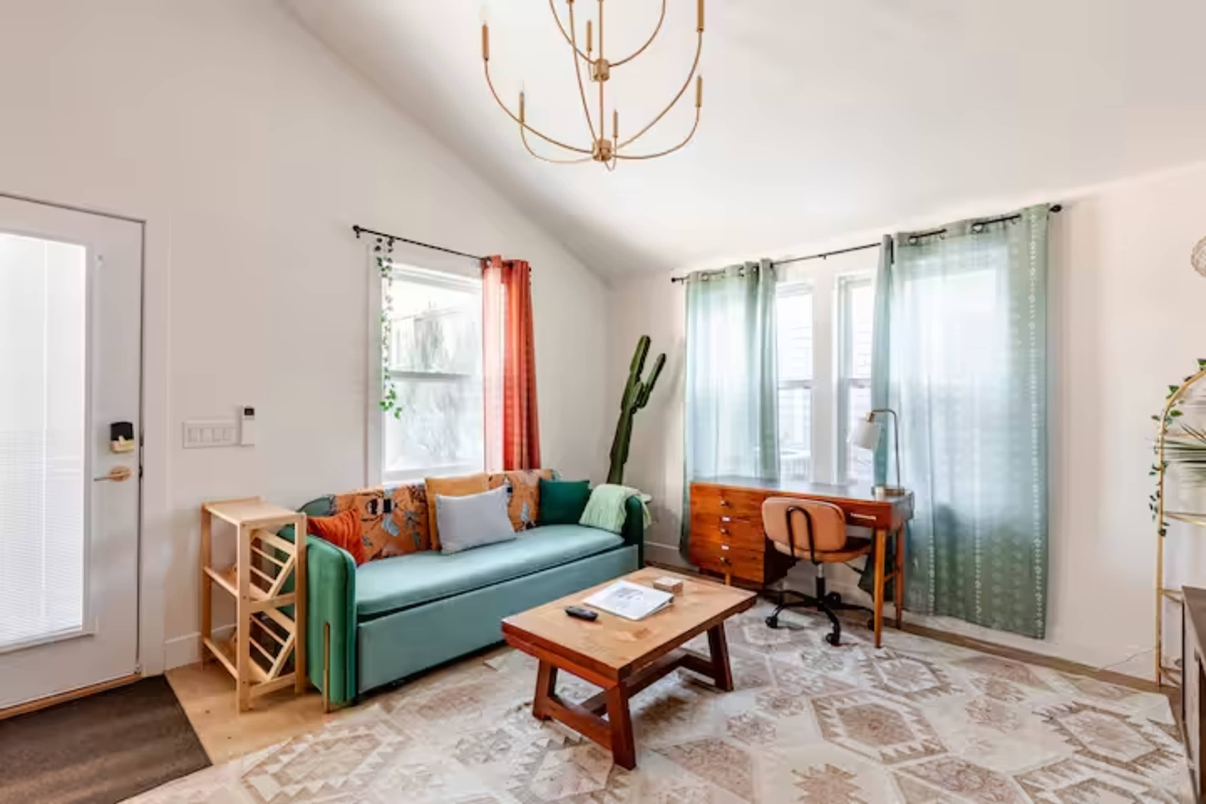 The image shows a cozy living room with a teal couch, a wooden coffee table, and a desk near a window, complemented by a cactus plant and decorative curtains.