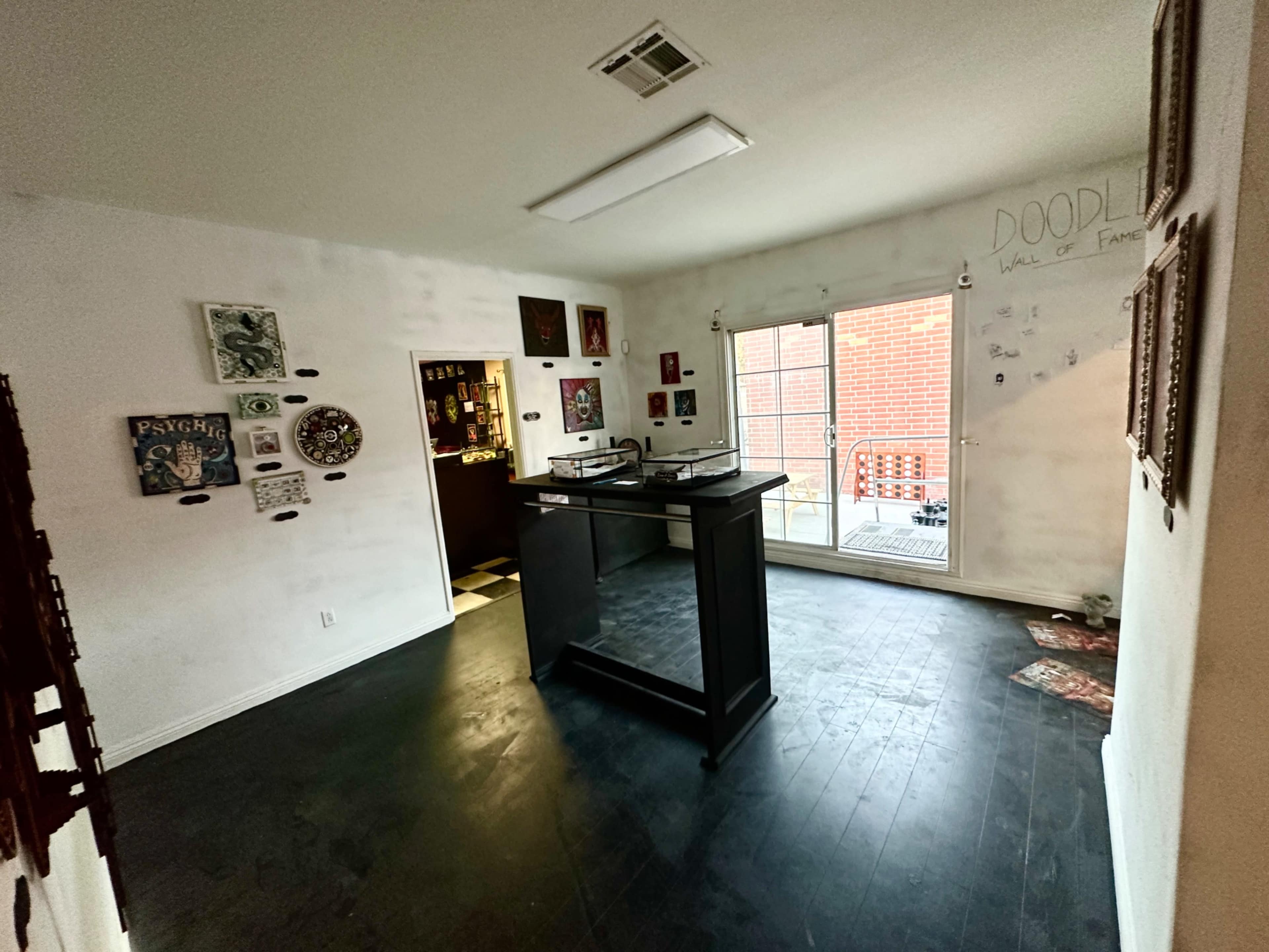 A spacious room with dark flooring features a central counter, walls adorned with various artworks, and a large window exposing an outdoor area.