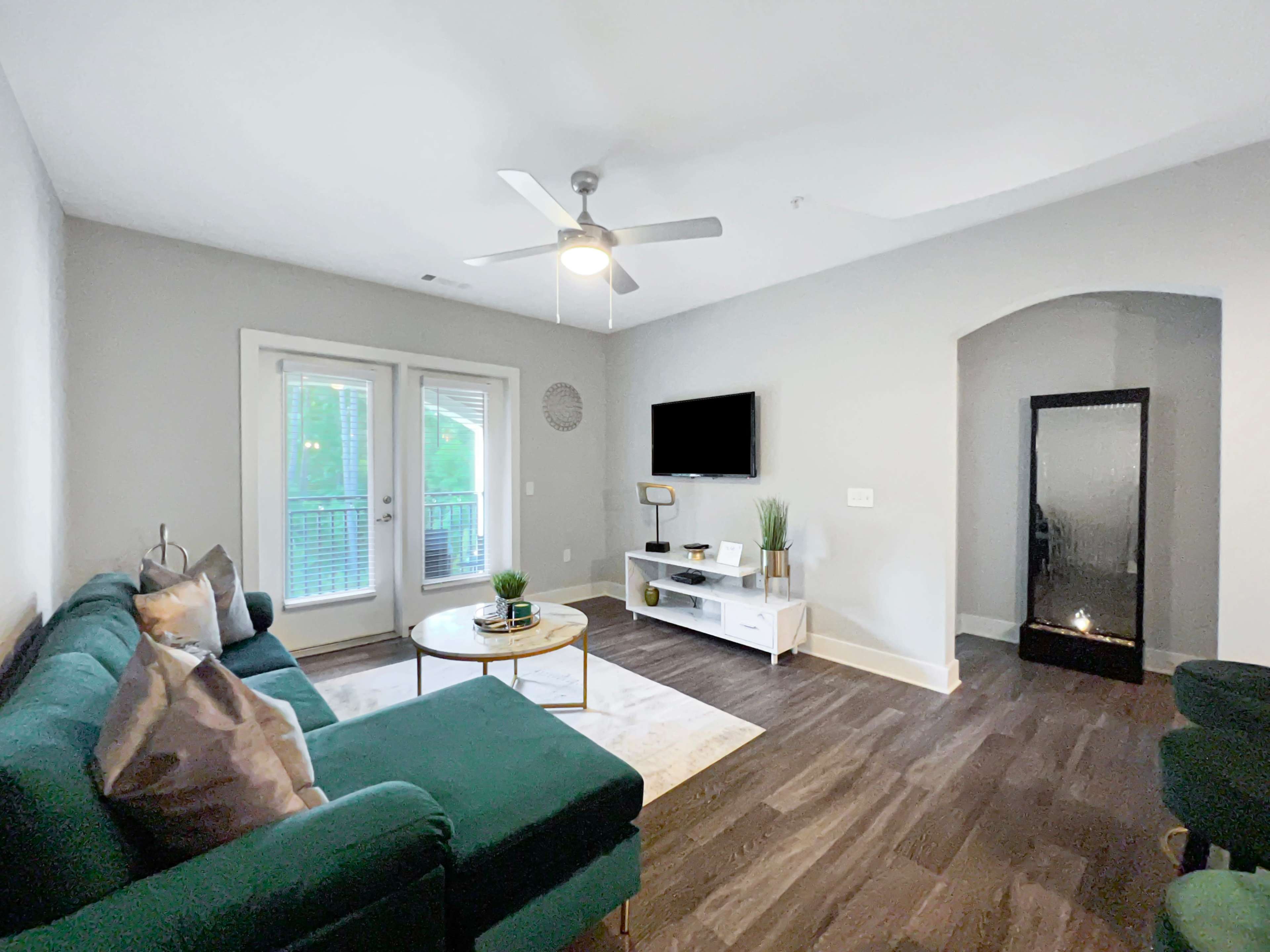 The living room features a green sofa, a round coffee table, a TV mounted on the wall, and large windows that open to a balcony.
