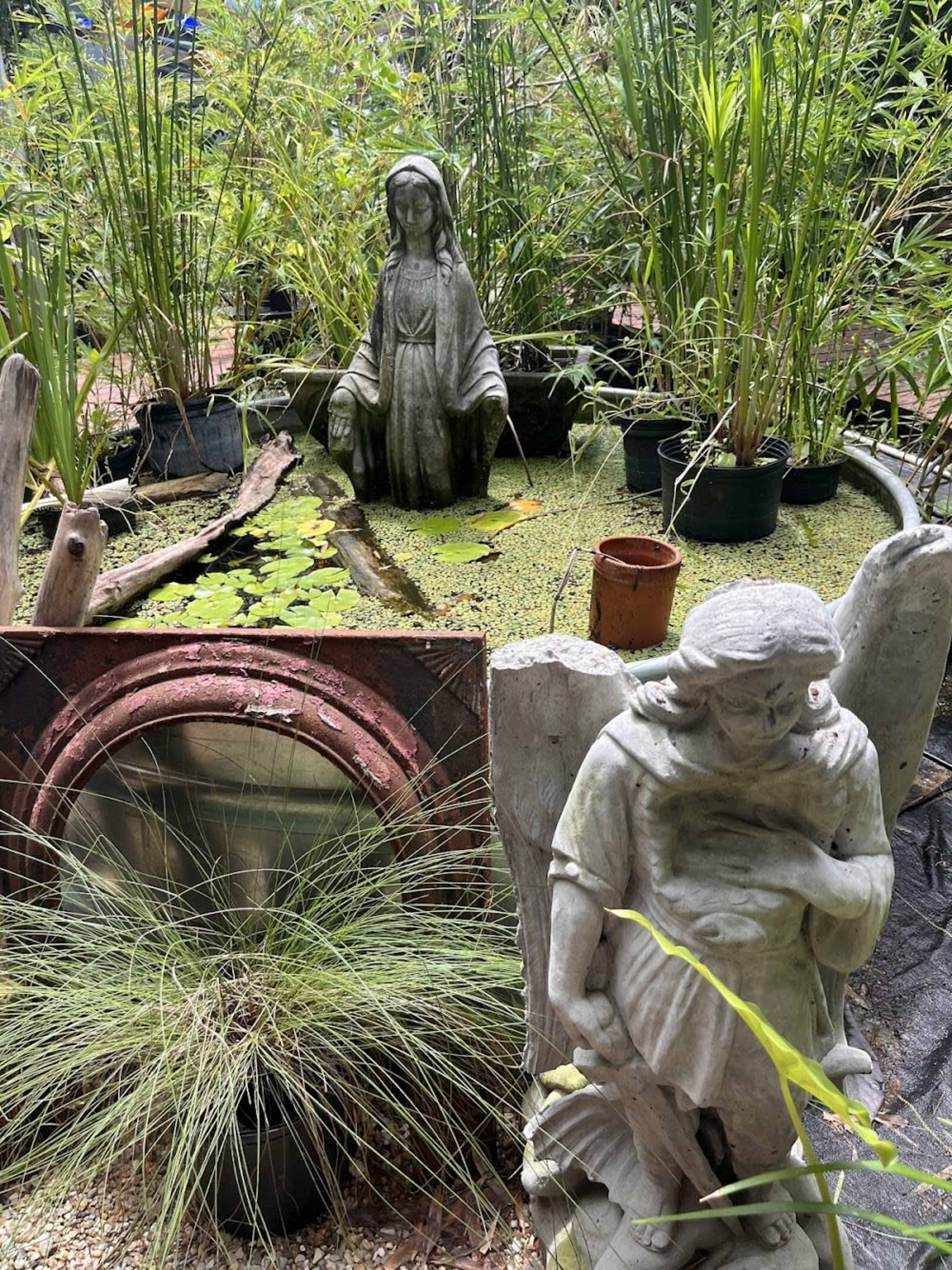The scene features two stone statues, one depicting a seated figure and the other an angel, surrounded by potted plants and a small pond covered in green vegetation.