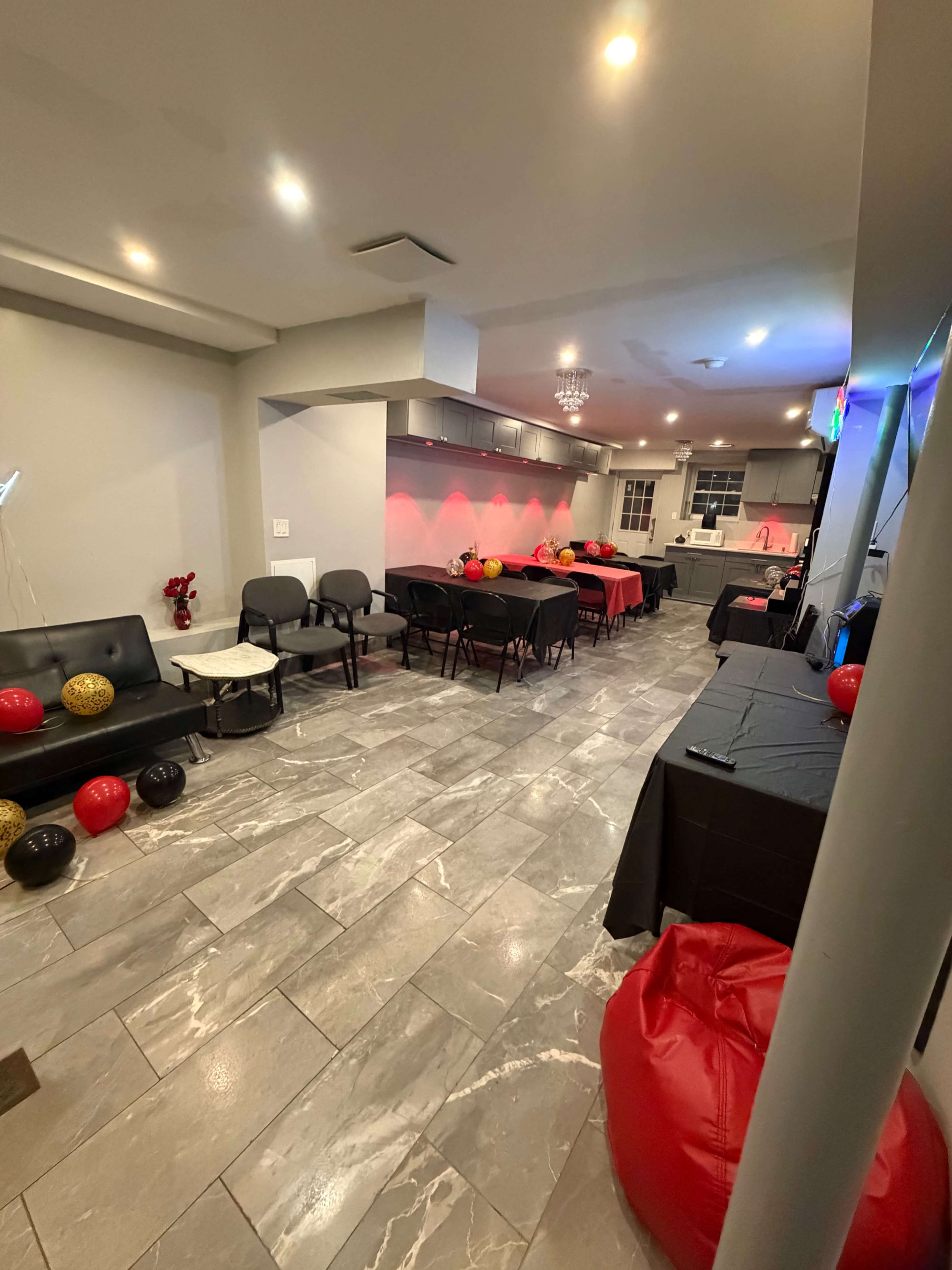 The image shows a party setup in a spacious room featuring black and red tables, chairs, and decorations, with balloons scattered throughout the area.