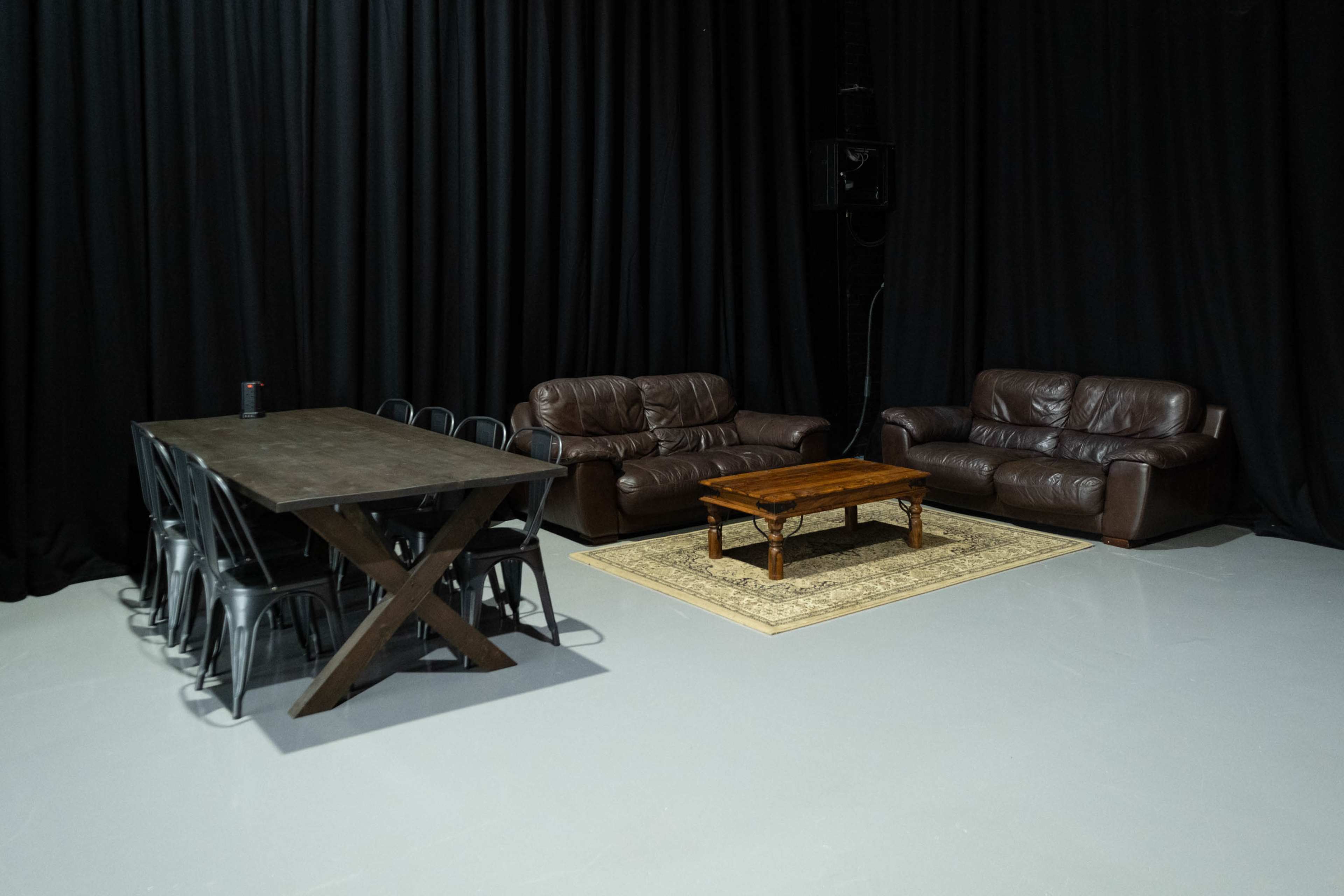 A black-curtained room features a wooden dining table with metal chairs on one side and two leather sofas with a coffee table and a rug on the other.